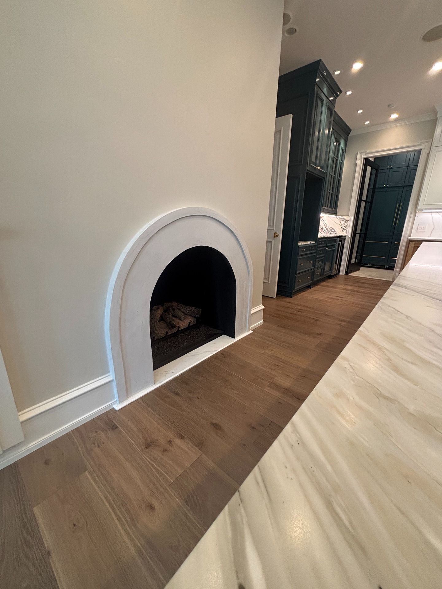 An arched fireplace set in a light-colored wall beside a wooden floor and a dark blue kitchen interior.