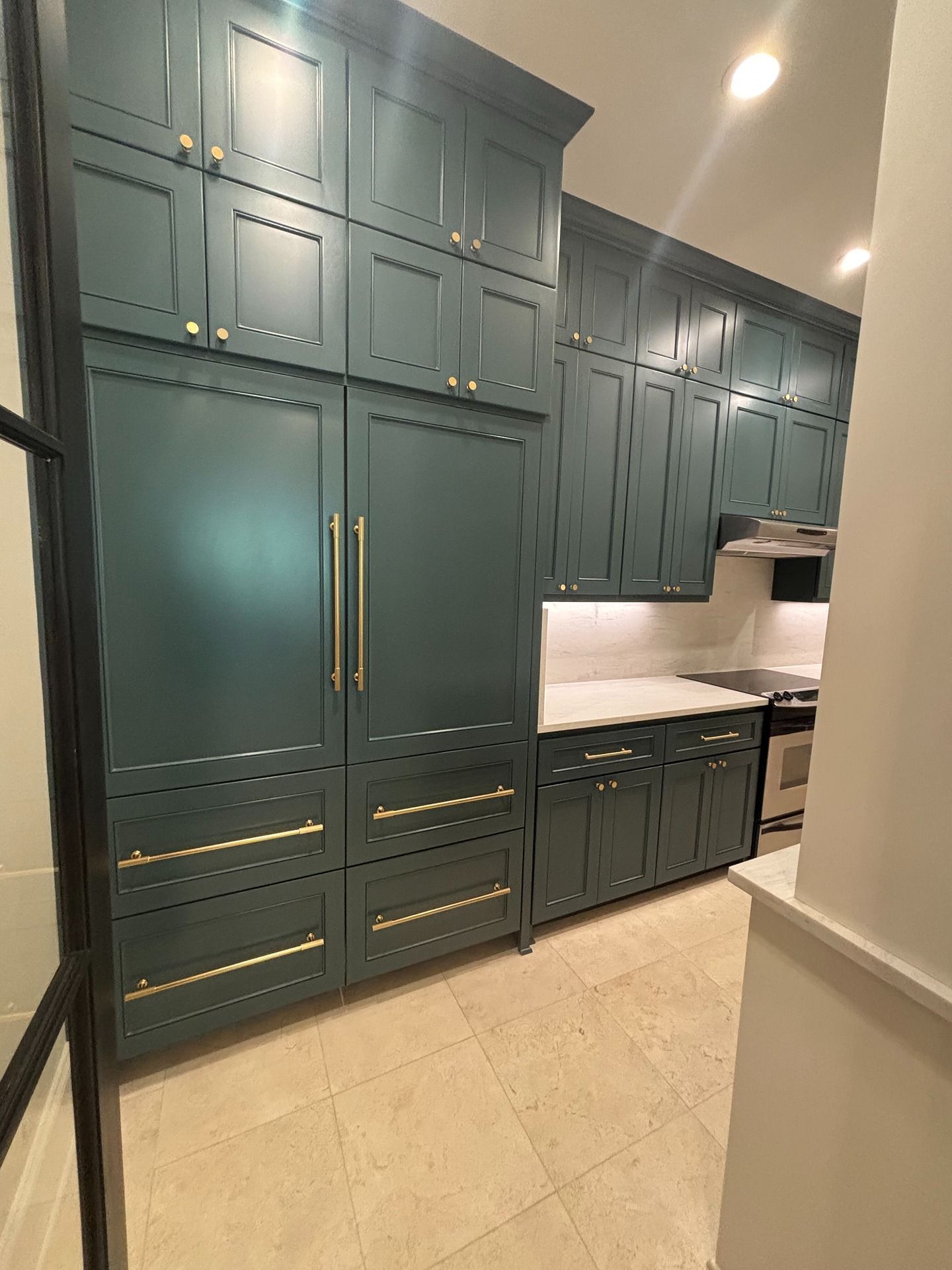 A modern kitchen featuring dark teal cabinetry with gold hardware, a built-in refrigerator, and stone tile flooring.