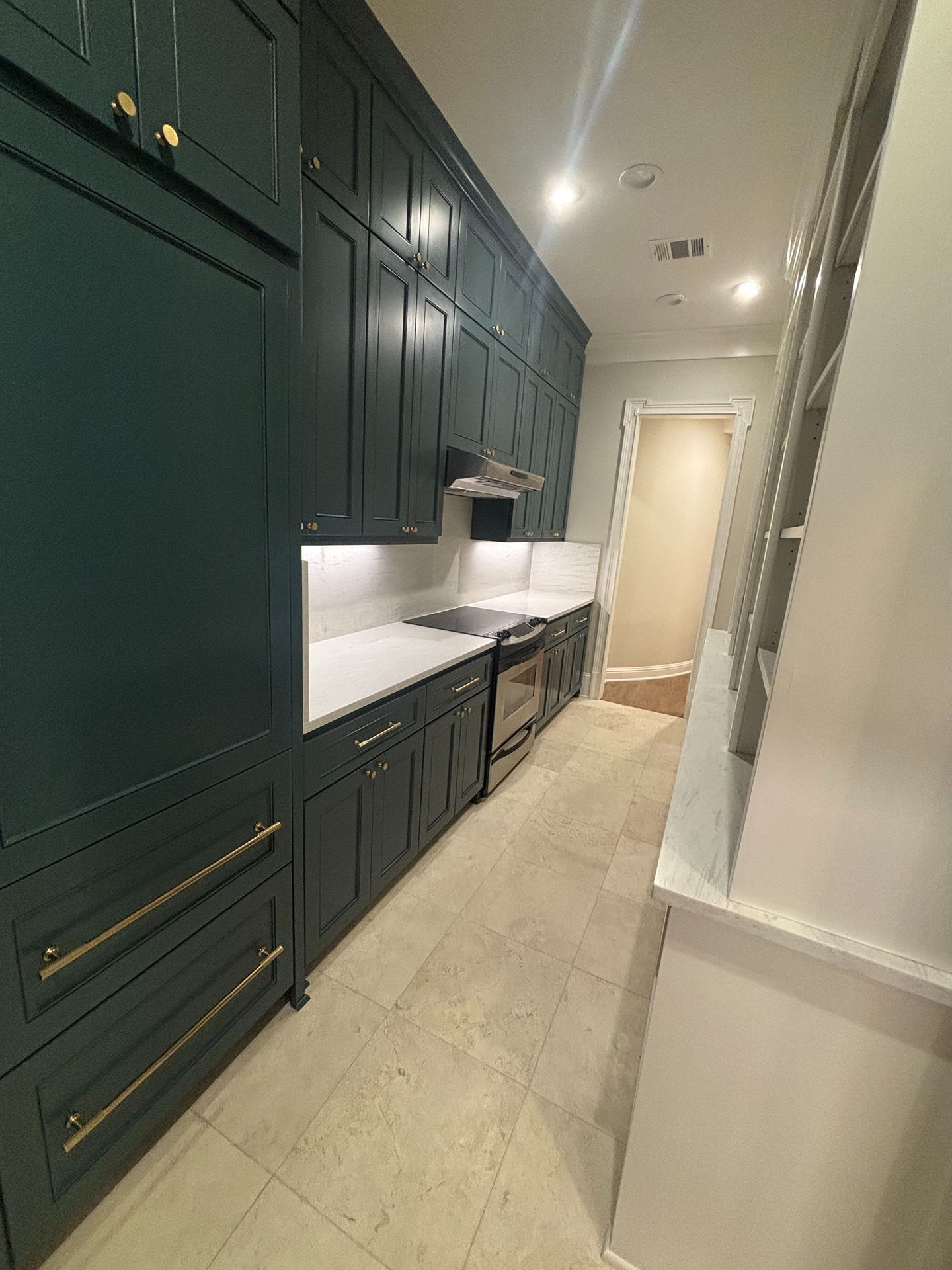 A galley-style kitchen with dark teal floor-to-ceiling cabinets, brass hardware, light countertops, and tiled flooring.