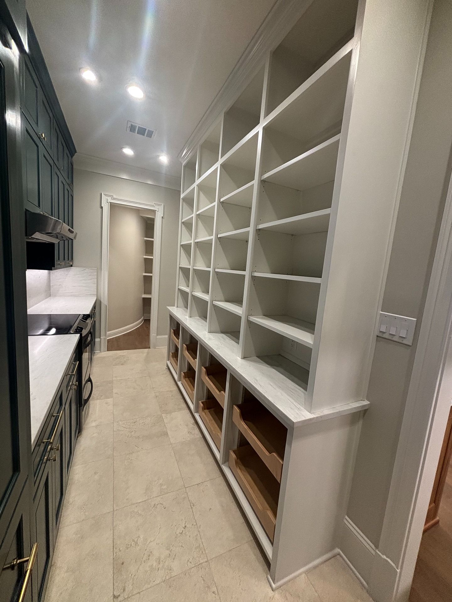 A modern butler's pantry with dark cabinets, white open shelving, drawers, and light-colored tile floors.