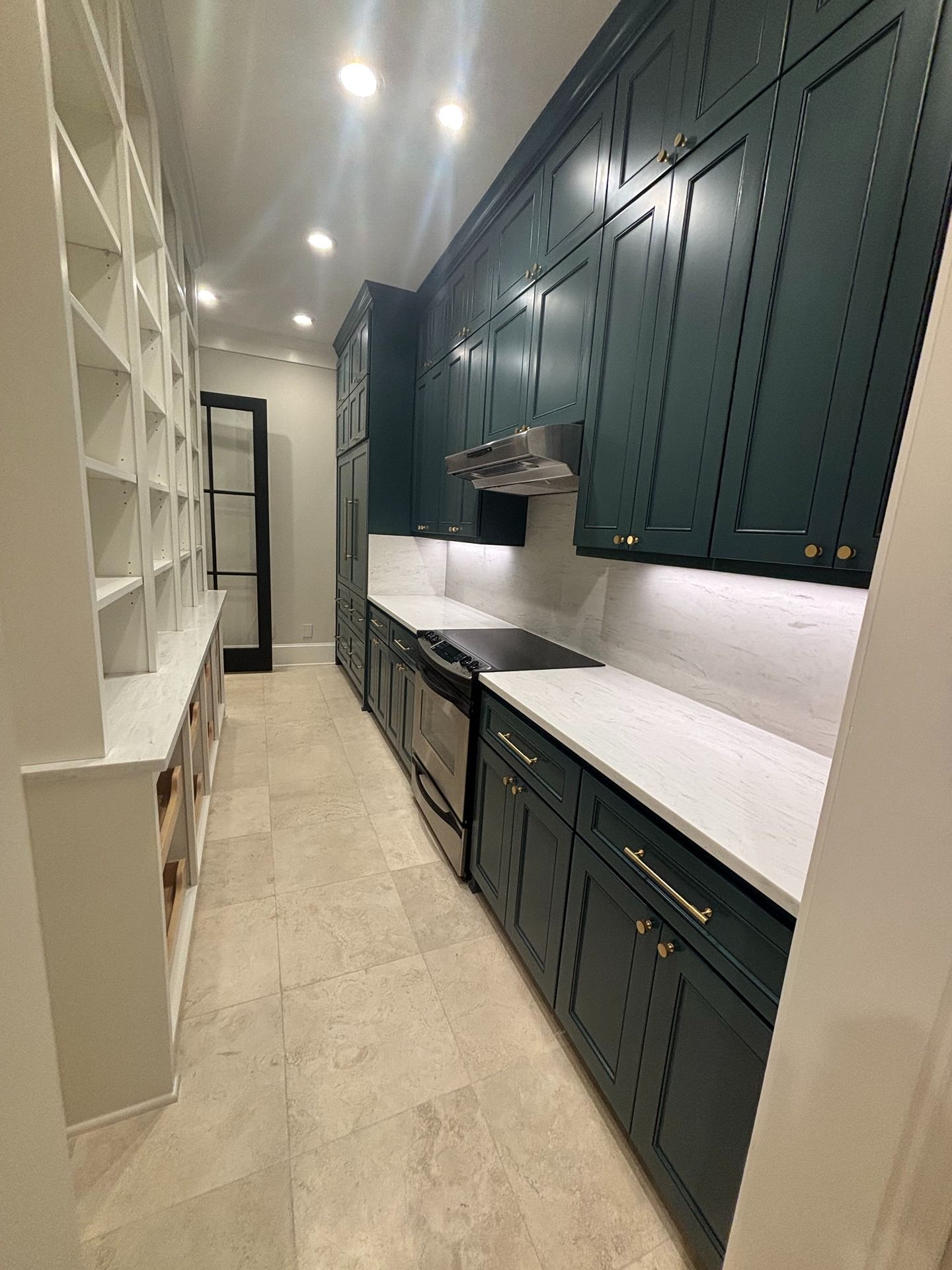 A long, galley-style kitchen pantry with teal cabinetry, white countertops, stone flooring, and built-in shelving.