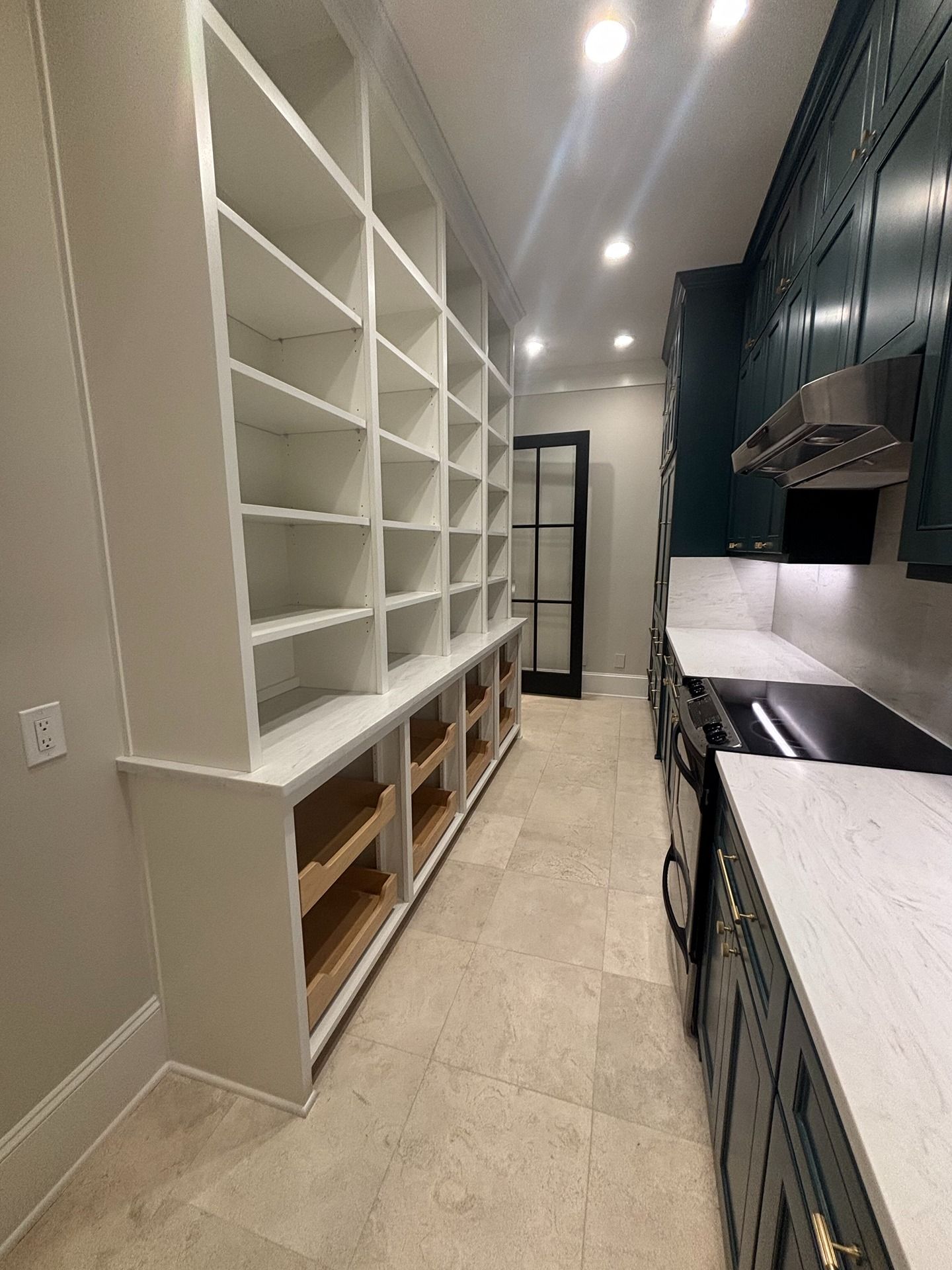 A narrow pantry features floor-to-ceiling white shelving on the left and dark green cabinets with stone counters on right.