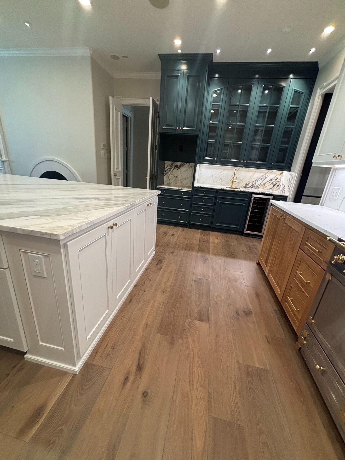 A kitchen with a large white island, dark green upper cabinets, wooden lower cabinets, and light wood flooring.