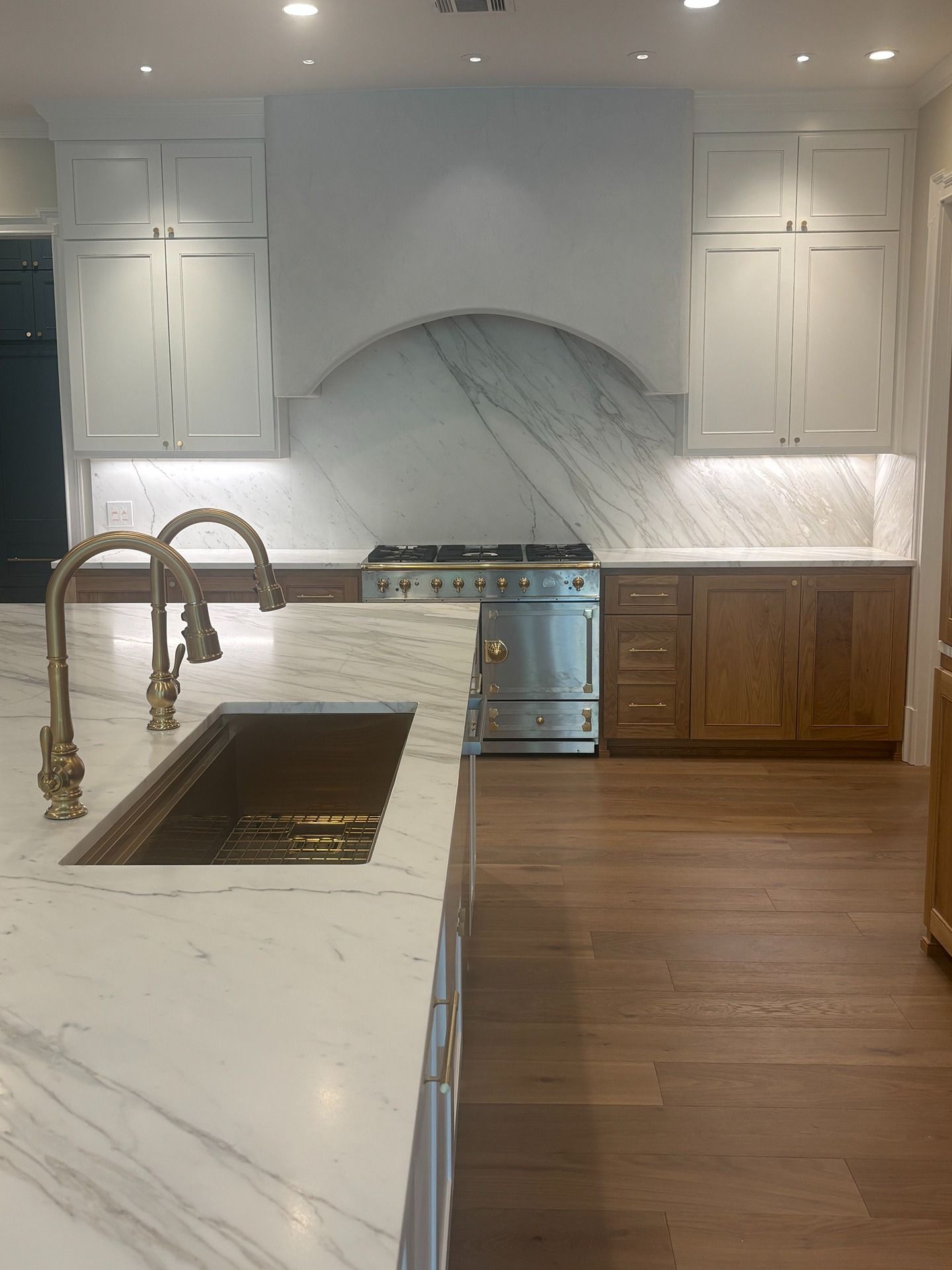Modern kitchen with white upper cabinets, wood lower cabinets, a marble island with a gold faucet, and a range with hood.