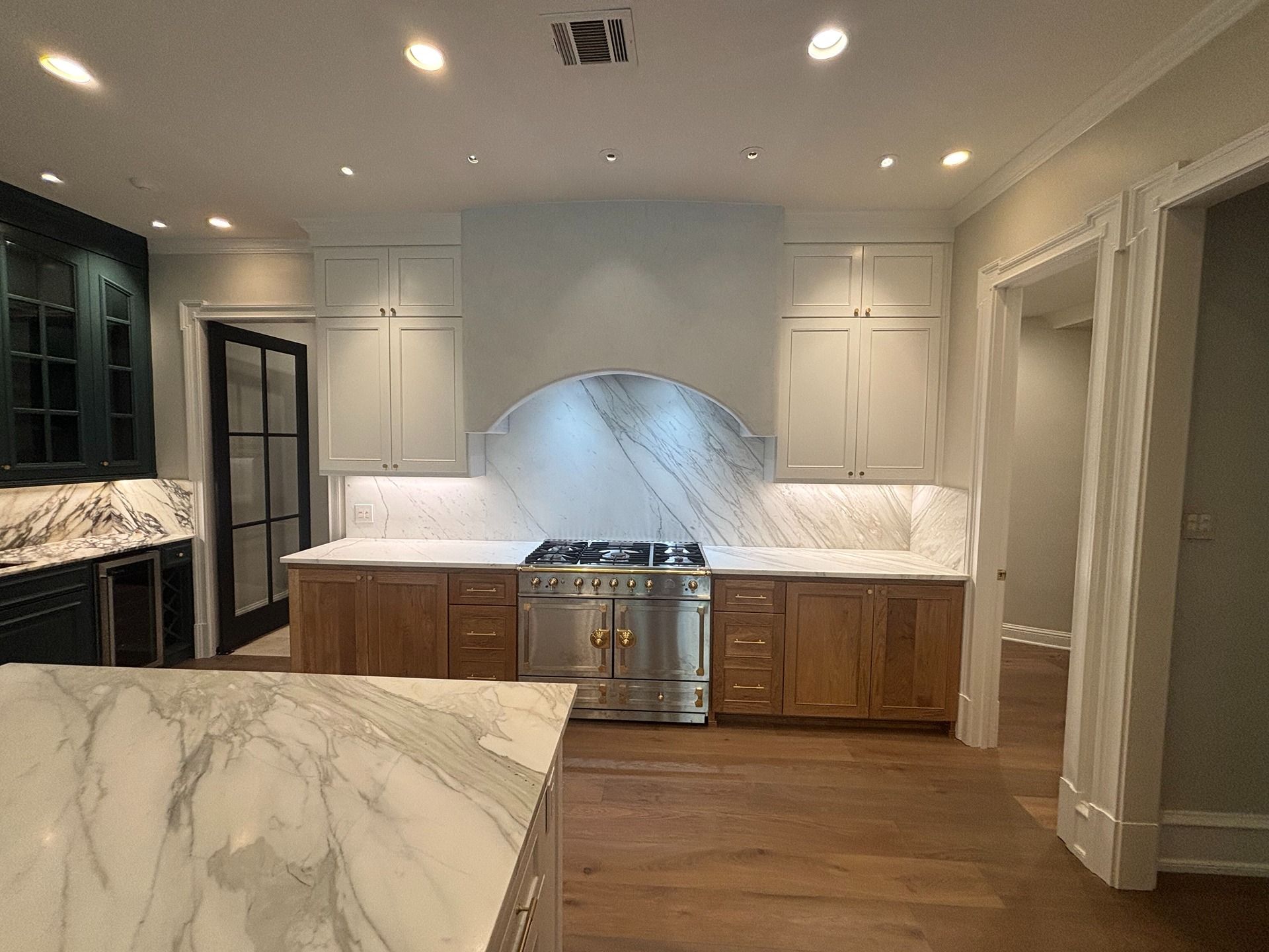 A modern kitchen with marble countertops, light wood cabinets, a range with a curved hood, and dark cabinetry on the left.