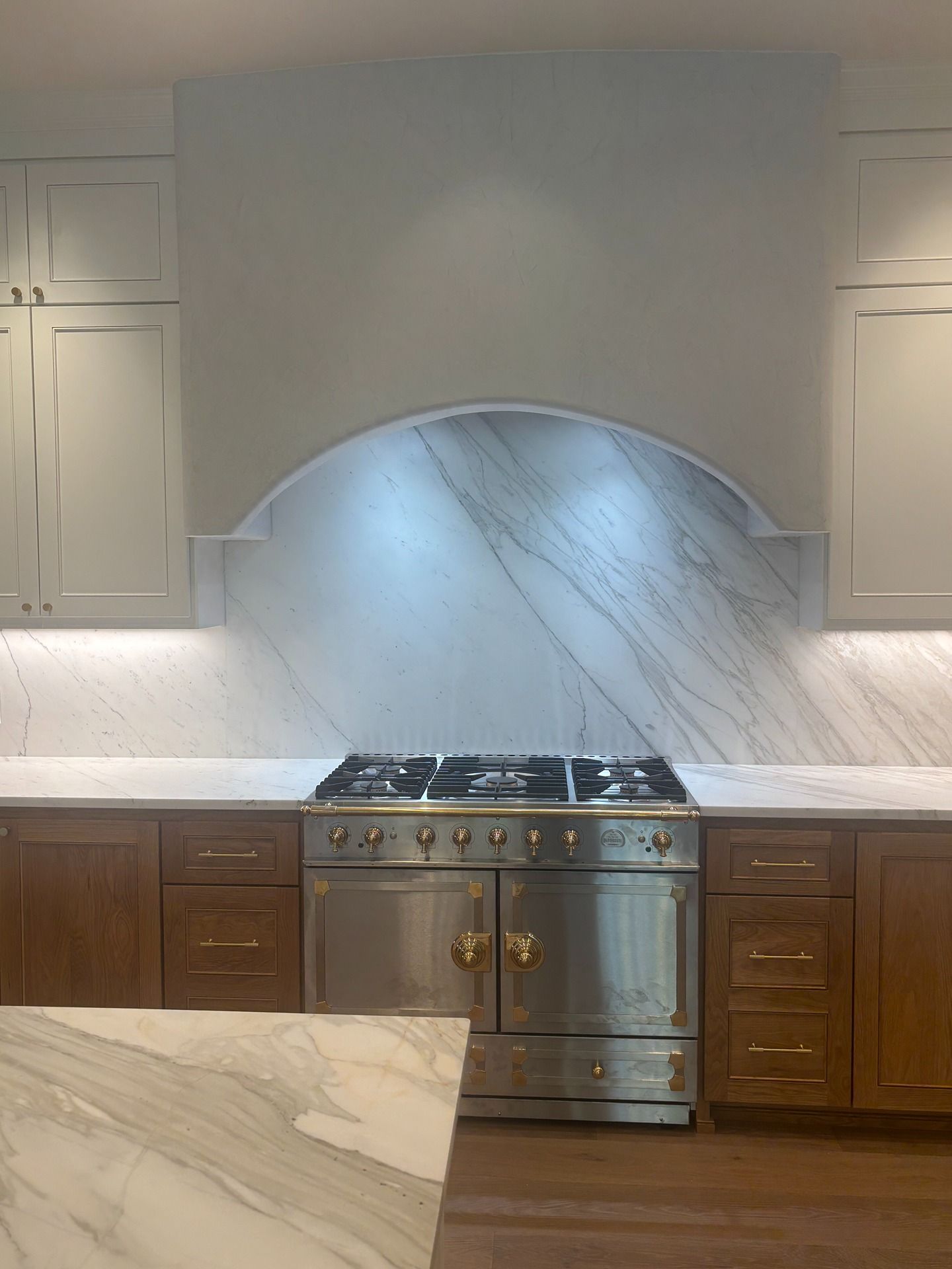 A kitchen featuring a stainless steel stove with a marble backsplash and custom range hood between wood and white cabinets.