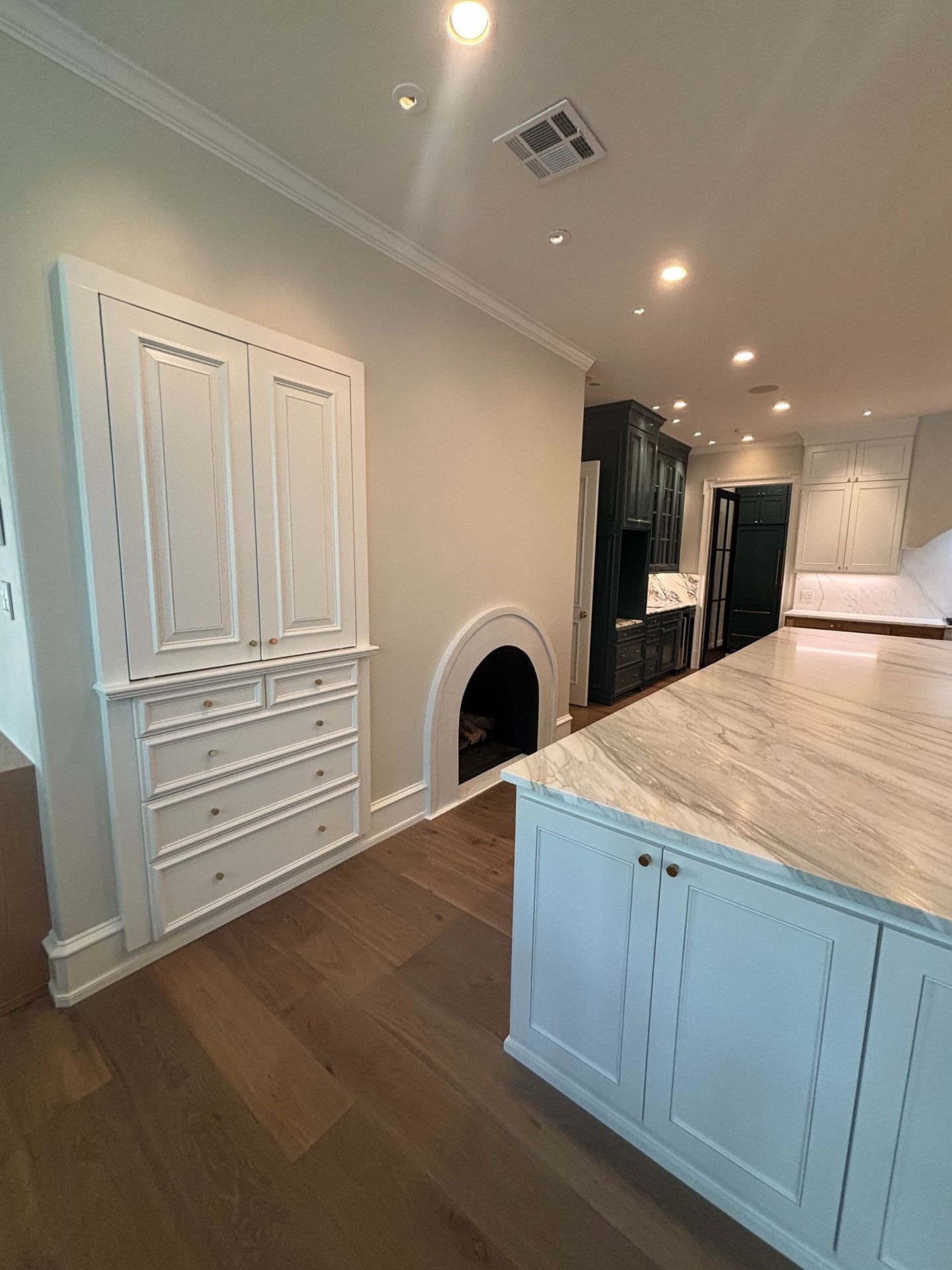 A kitchen features a white built-in cabinet and arched hearth beside a large marble-topped island on hardwood floors.