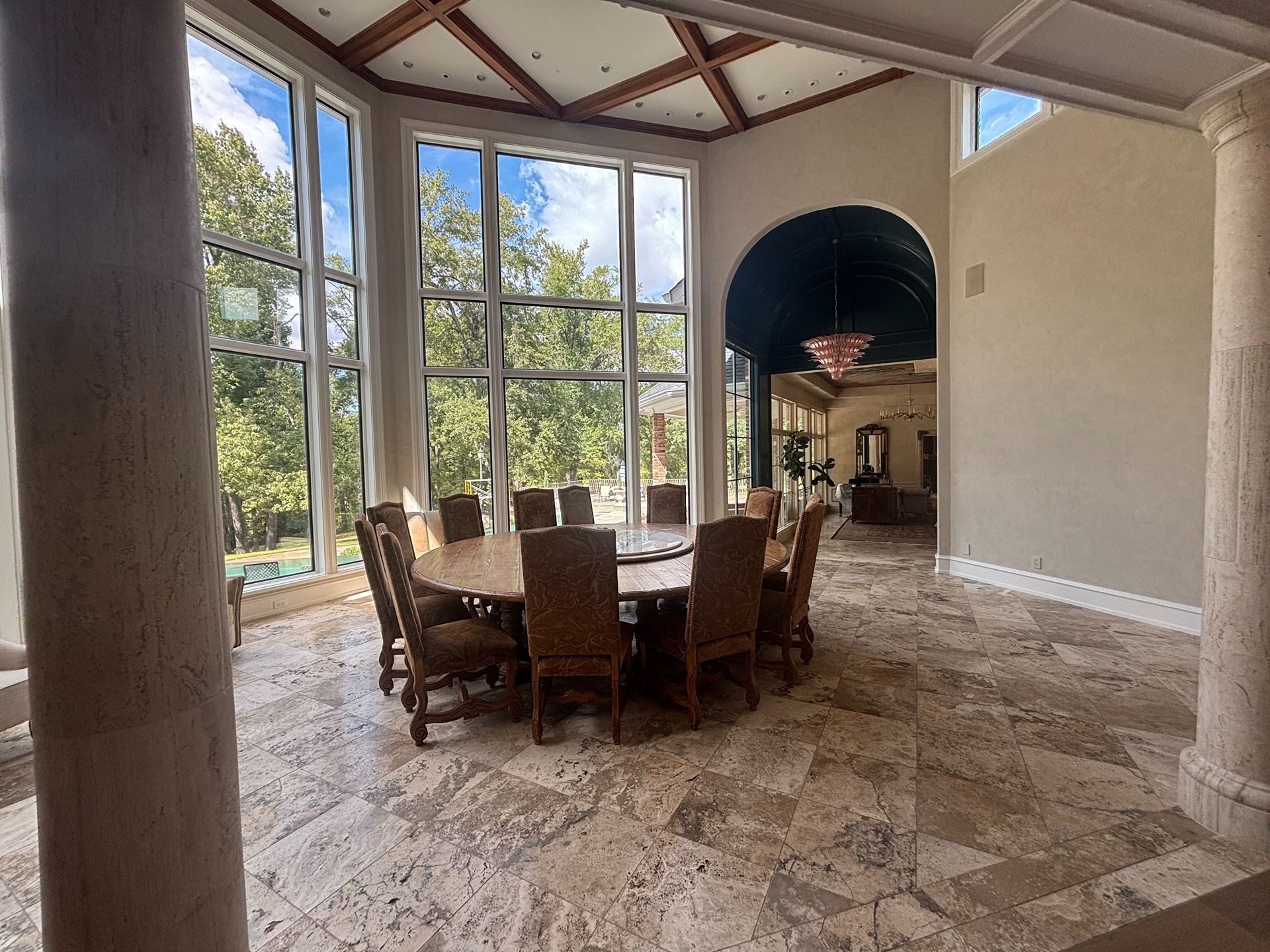 A large circular dining table with chairs in a sunlit room featuring tall windows, stone tile floors, and arched ceilings.