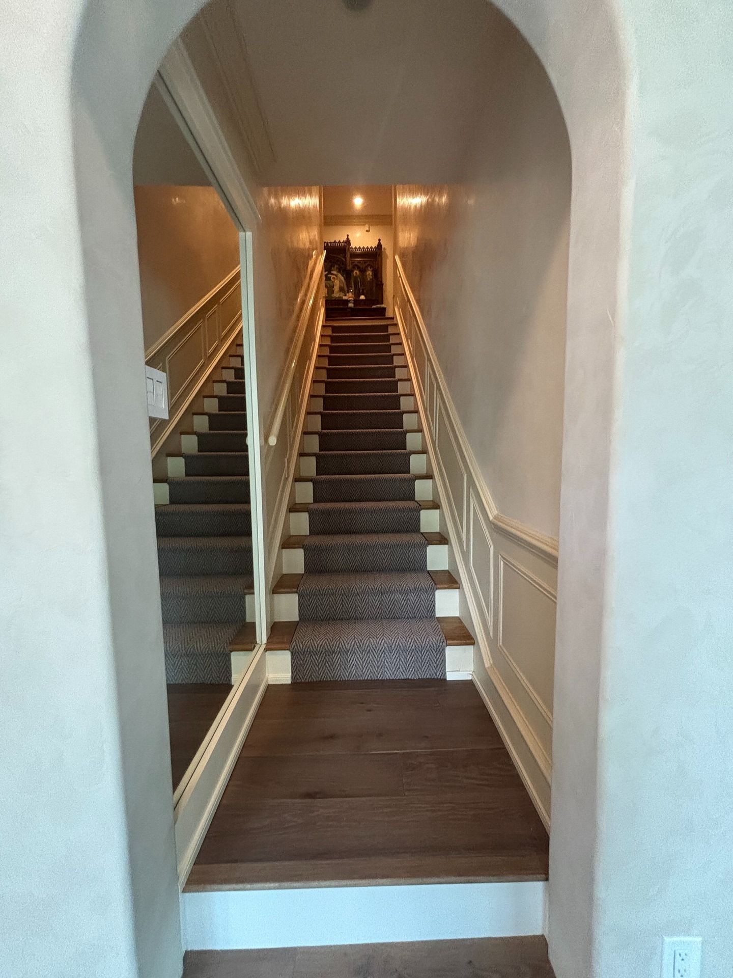 An arched entry leads to a straight staircase with a patterned carpet runner, framed by a wall mirror and wainscoting.