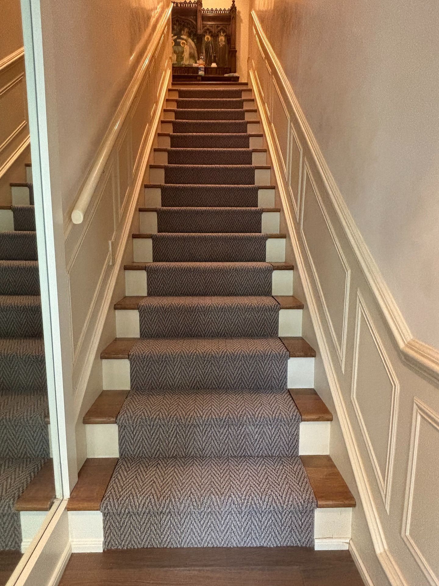 A staircase with wooden treads and a dark grey herringbone runner, bordered by white walls with molding and a mirror.