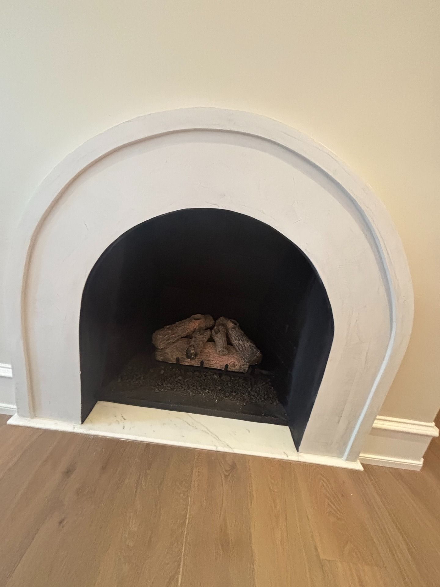 An arched fireplace with a white surround, dark interior, and a cluster of artificial logs set on a light wood floor.
