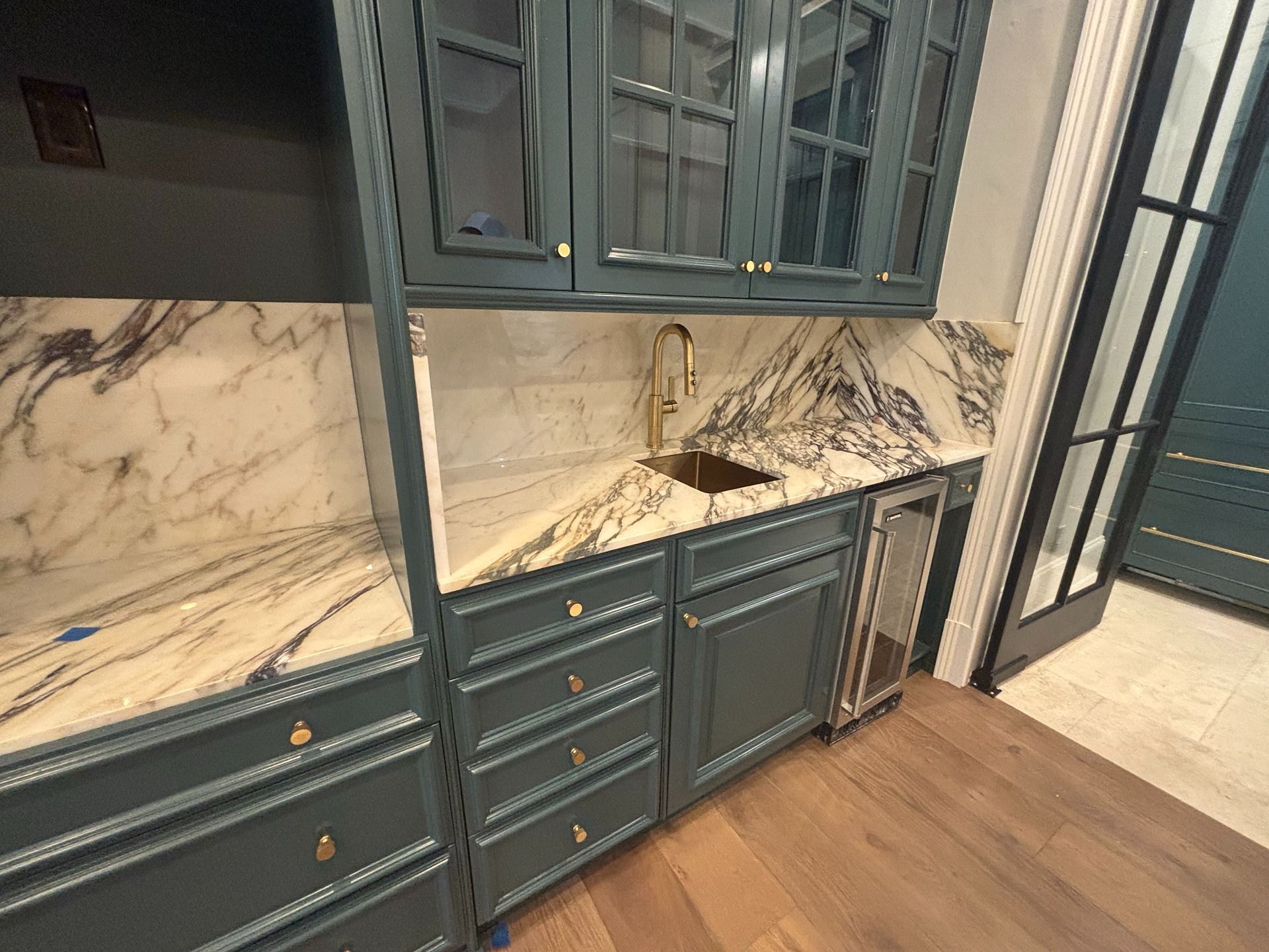 A modern wet bar featuring teal cabinets, a gold faucet, white marble countertops with dark veining, and wood flooring.