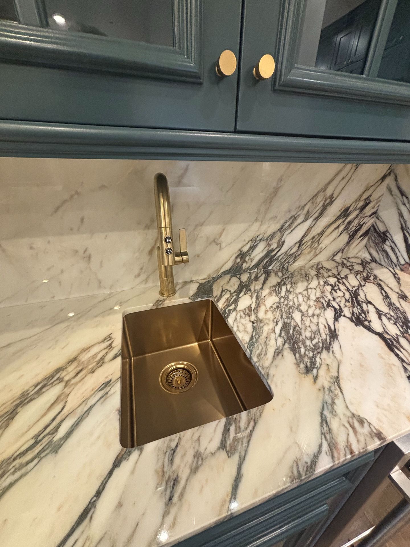 A gold sink and matching faucet set into a white and grey marble countertop beneath dark blue cabinets.
