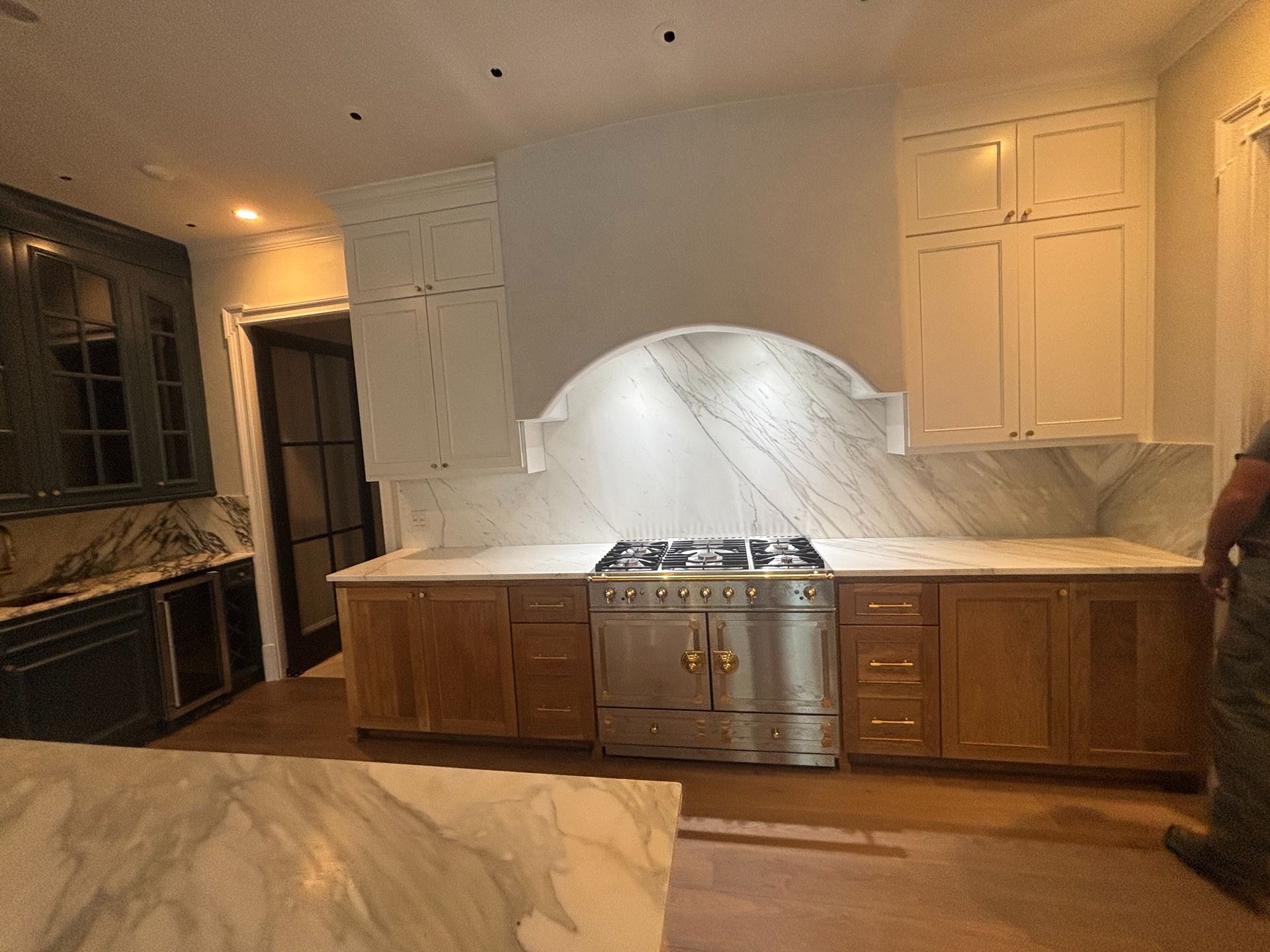 A kitchen features a range with a white arched hood, marble backsplash, wood cabinets, and a marble-topped island.