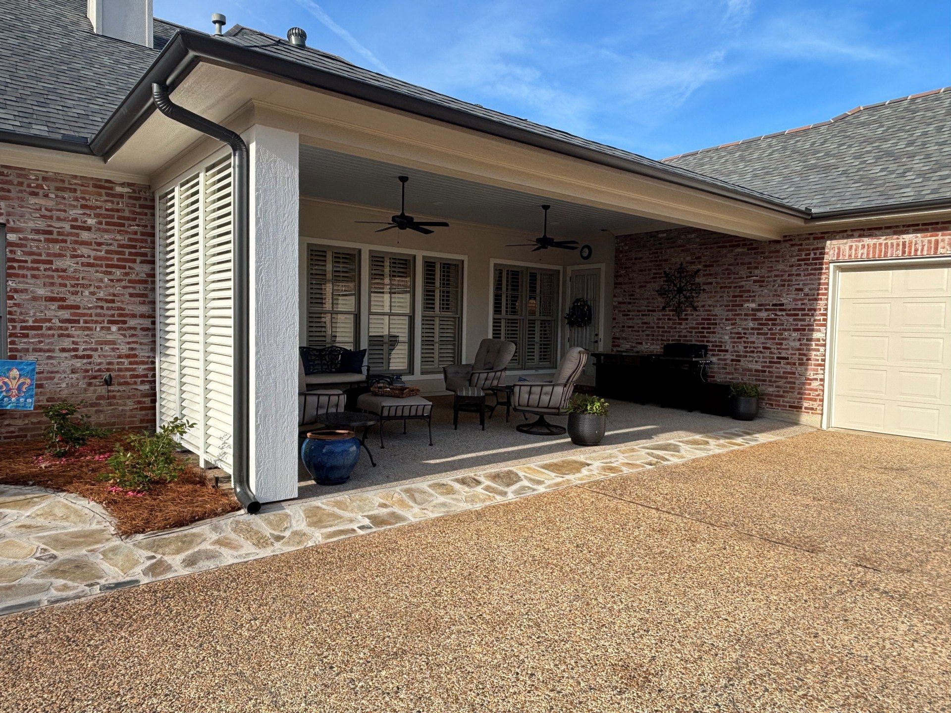 A covered brick patio with white louvered privacy screens, outdoor seating, ceiling fans, and a stone walkway.