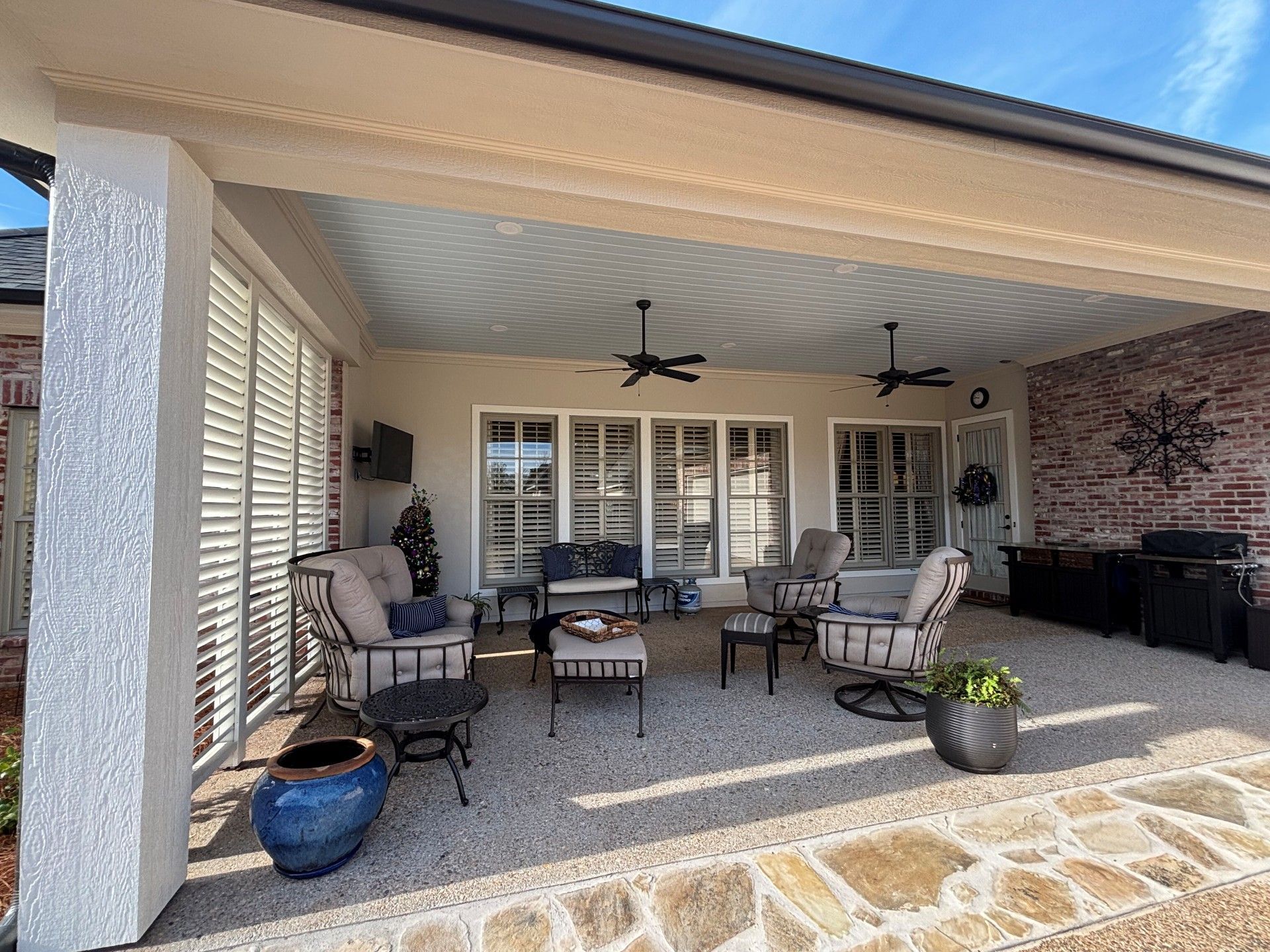 A covered patio featuring outdoor lounge chairs, a small table, ceiling fans, and a stone floor under a bright sky.