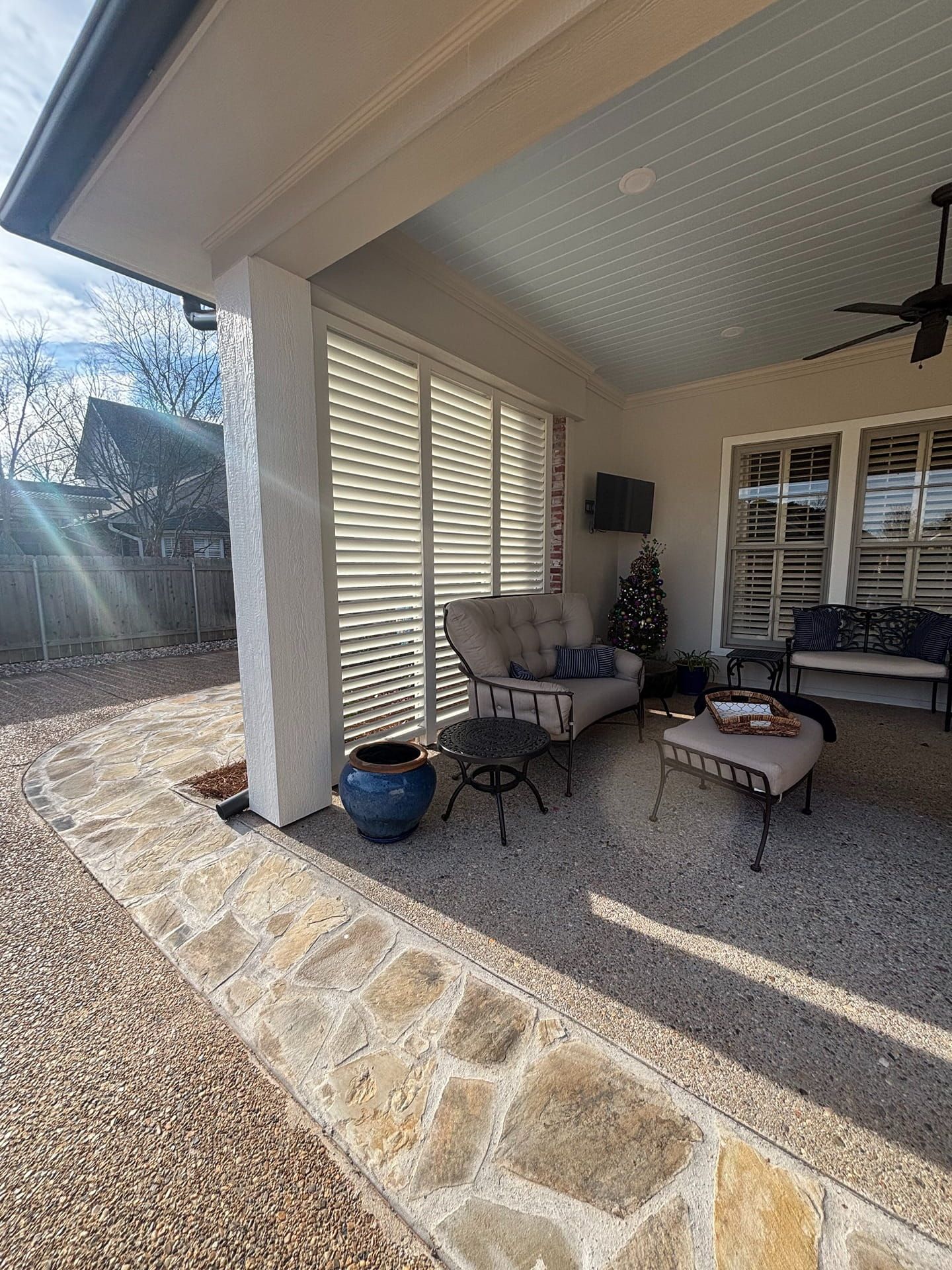 A covered patio featuring outdoor furniture, a blue planter, and white shutters on a stone floor with a gravel yard.