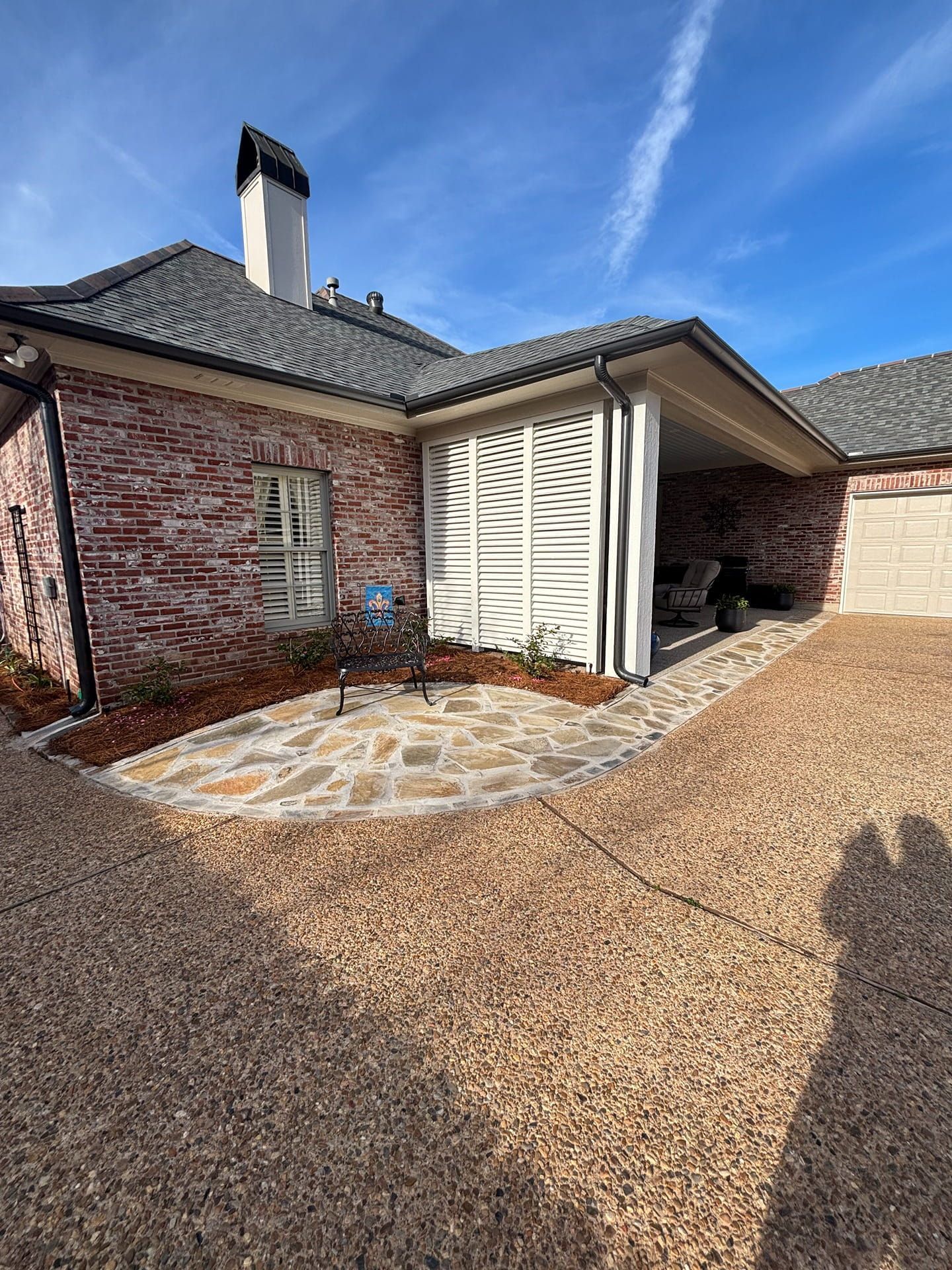 Red brick house with a white-slatted porch, stone walkway, and gravel driveway under a bright blue sky.