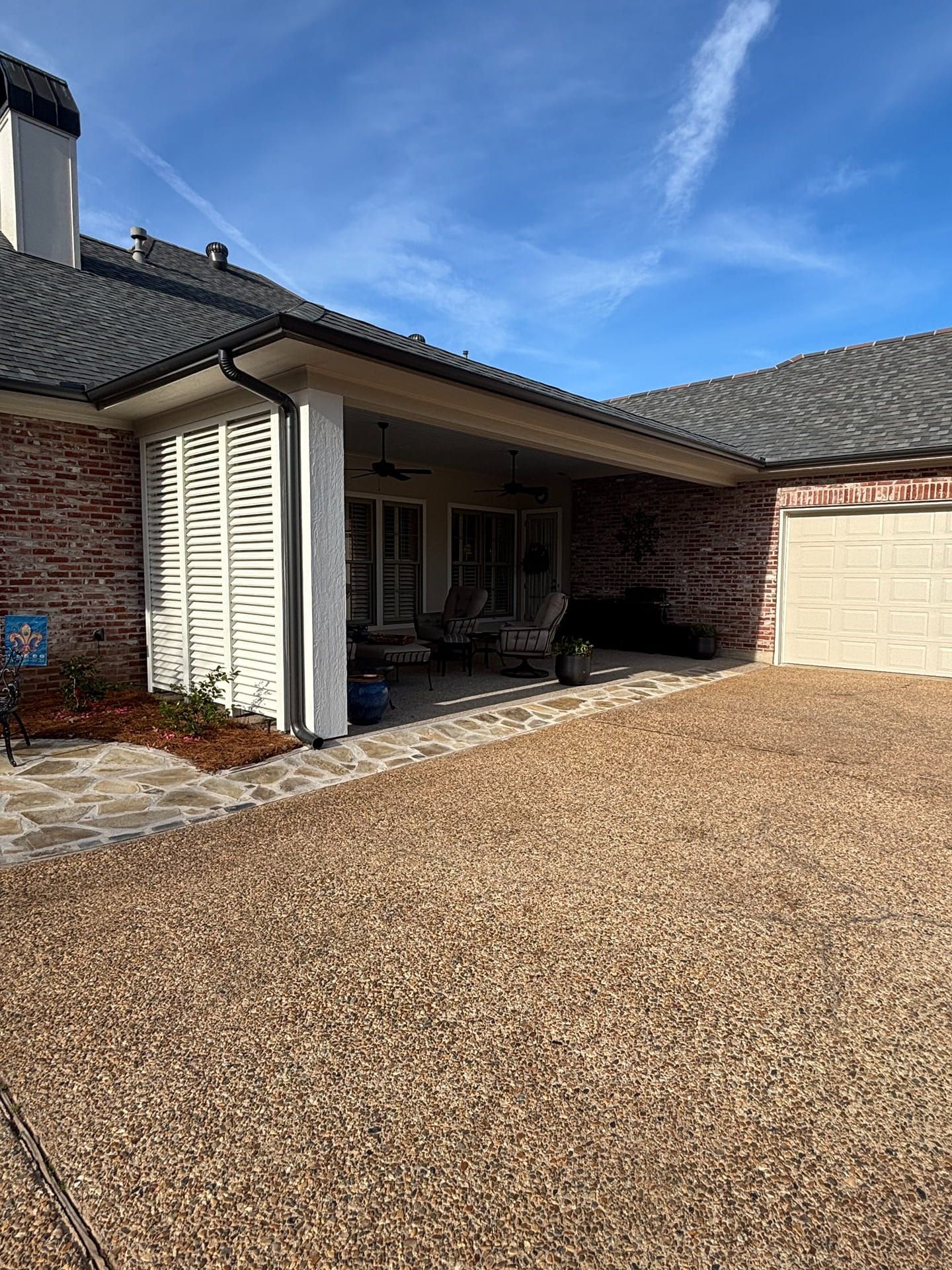 A brick house with a covered patio, louvered privacy screen, stone walkway, and a gravel driveway under a blue sky.
