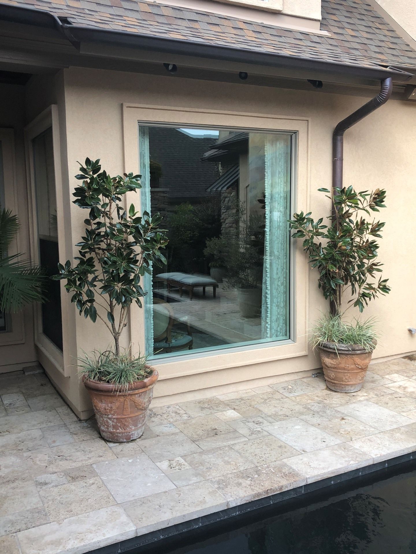 A beige wall features a large window reflecting a patio, flanked by two potted trees on a stone pool deck.