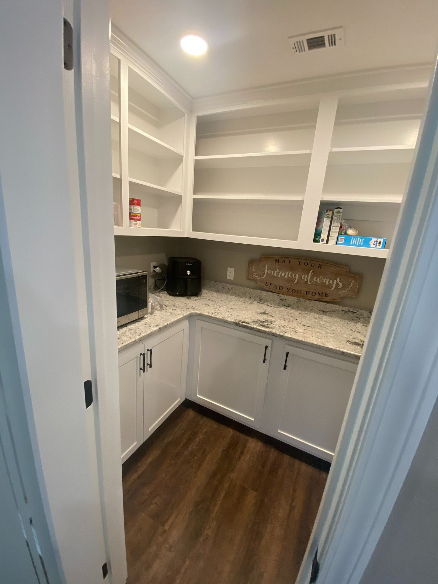 A white pantry with open shelving, granite countertops, lower cabinets, and dark wood flooring.