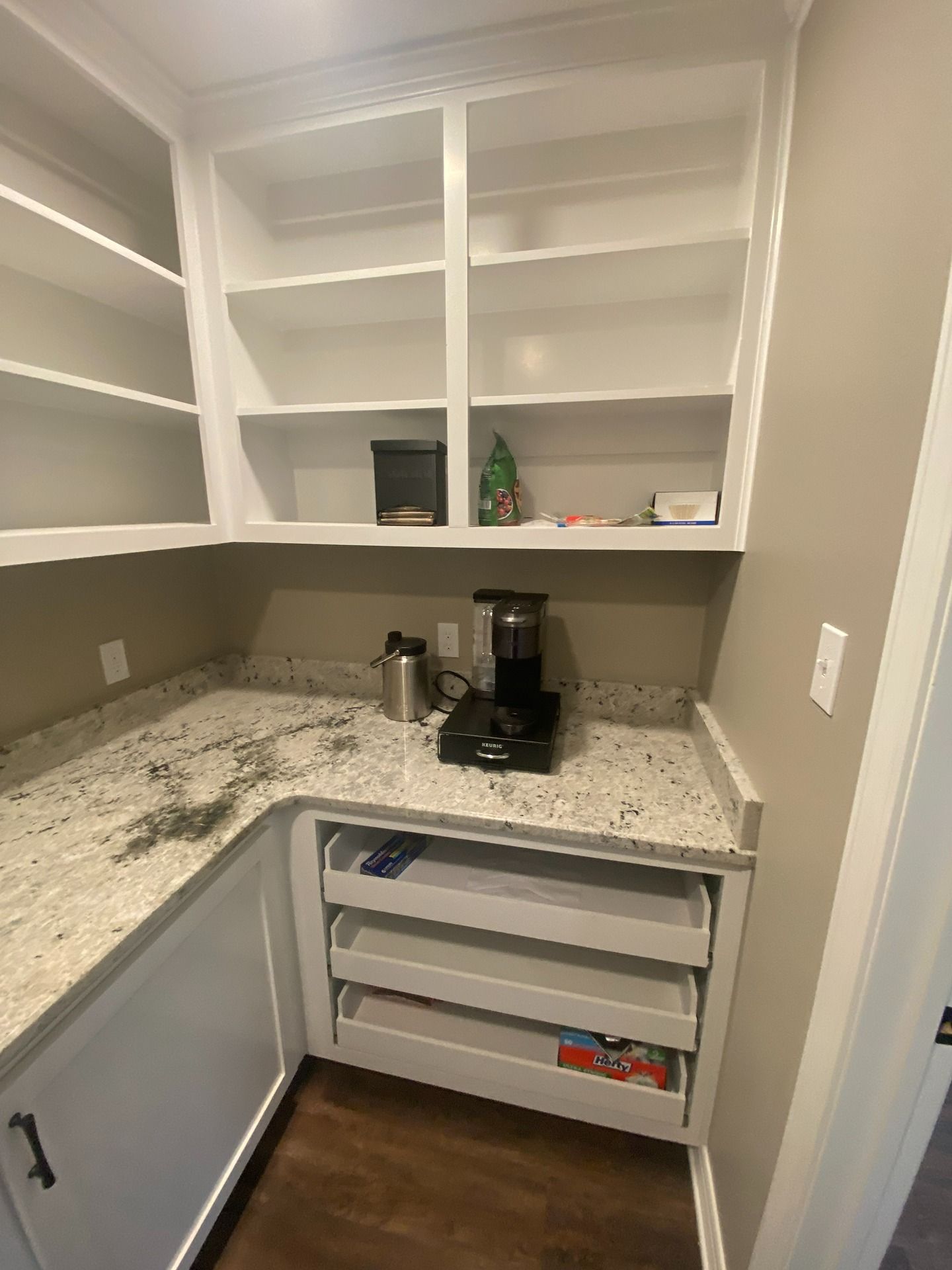 A kitchen pantry with white cabinets, speckled granite countertops, open shelving, and three pull-out storage drawers.