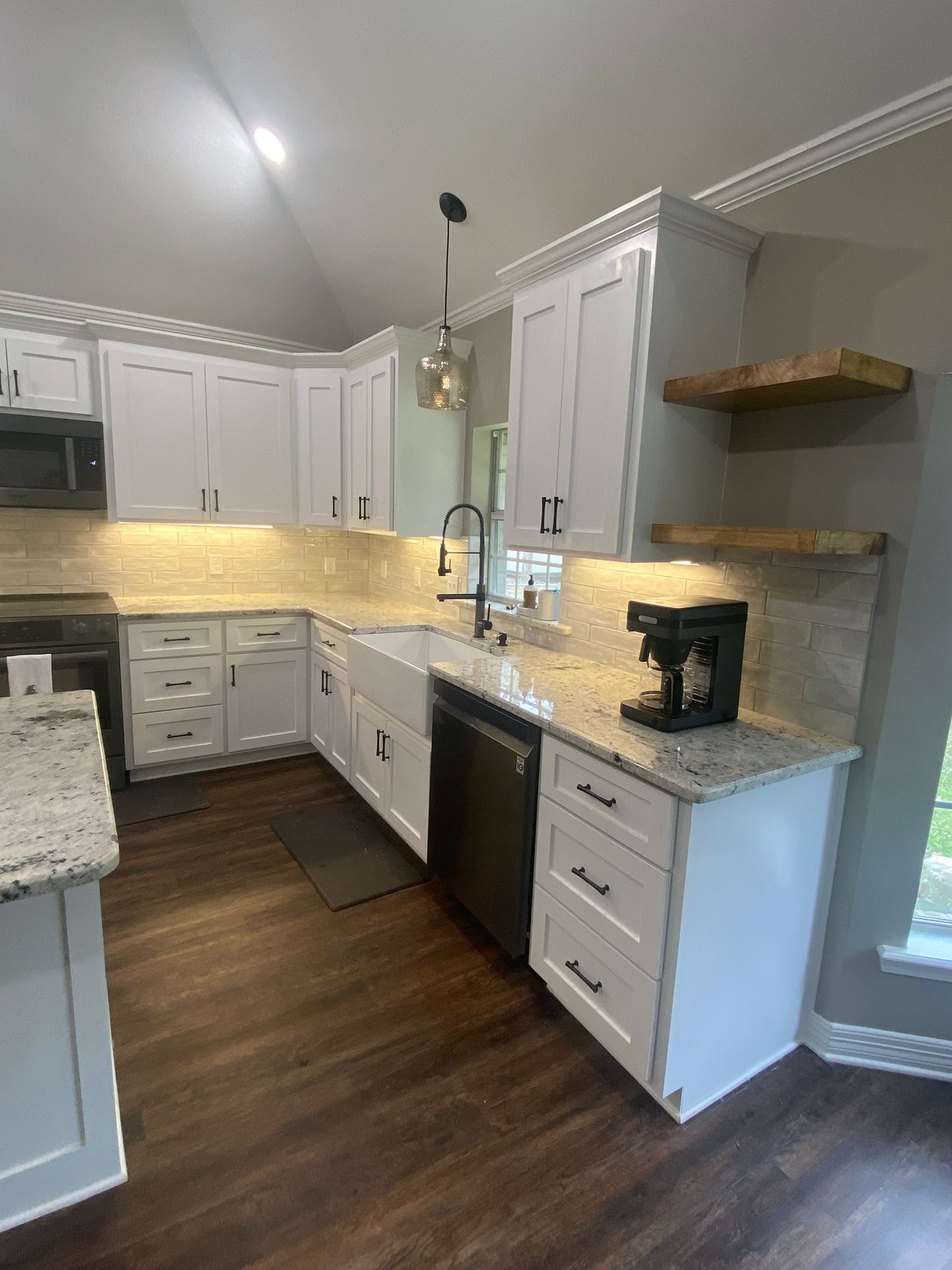 A bright kitchen with white cabinets, light granite countertops, a farmhouse sink, and two rustic wood floating shelves.