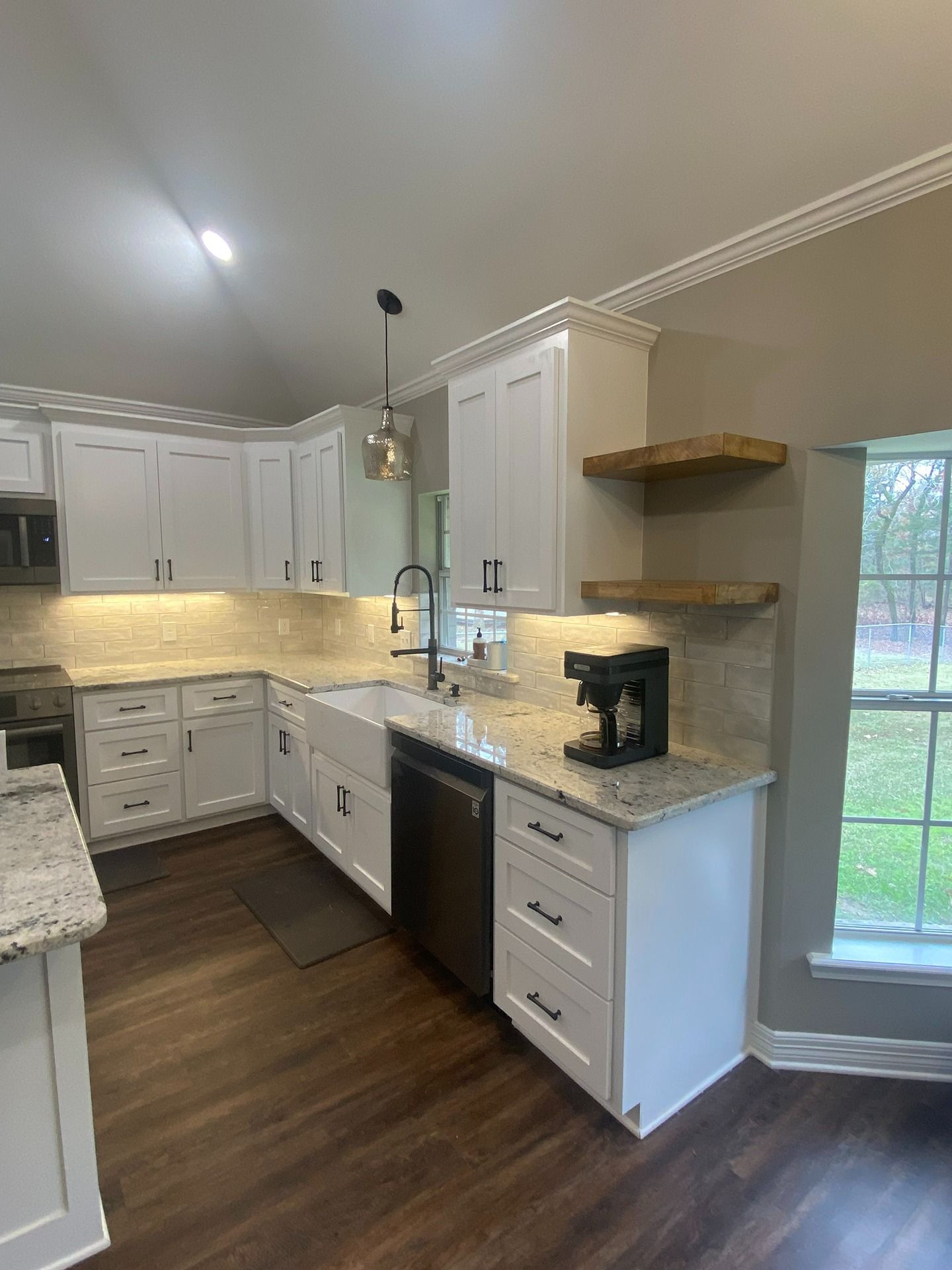 A bright, modern kitchen featuring white cabinets, granite countertops, wood open shelving, and dark wood flooring.