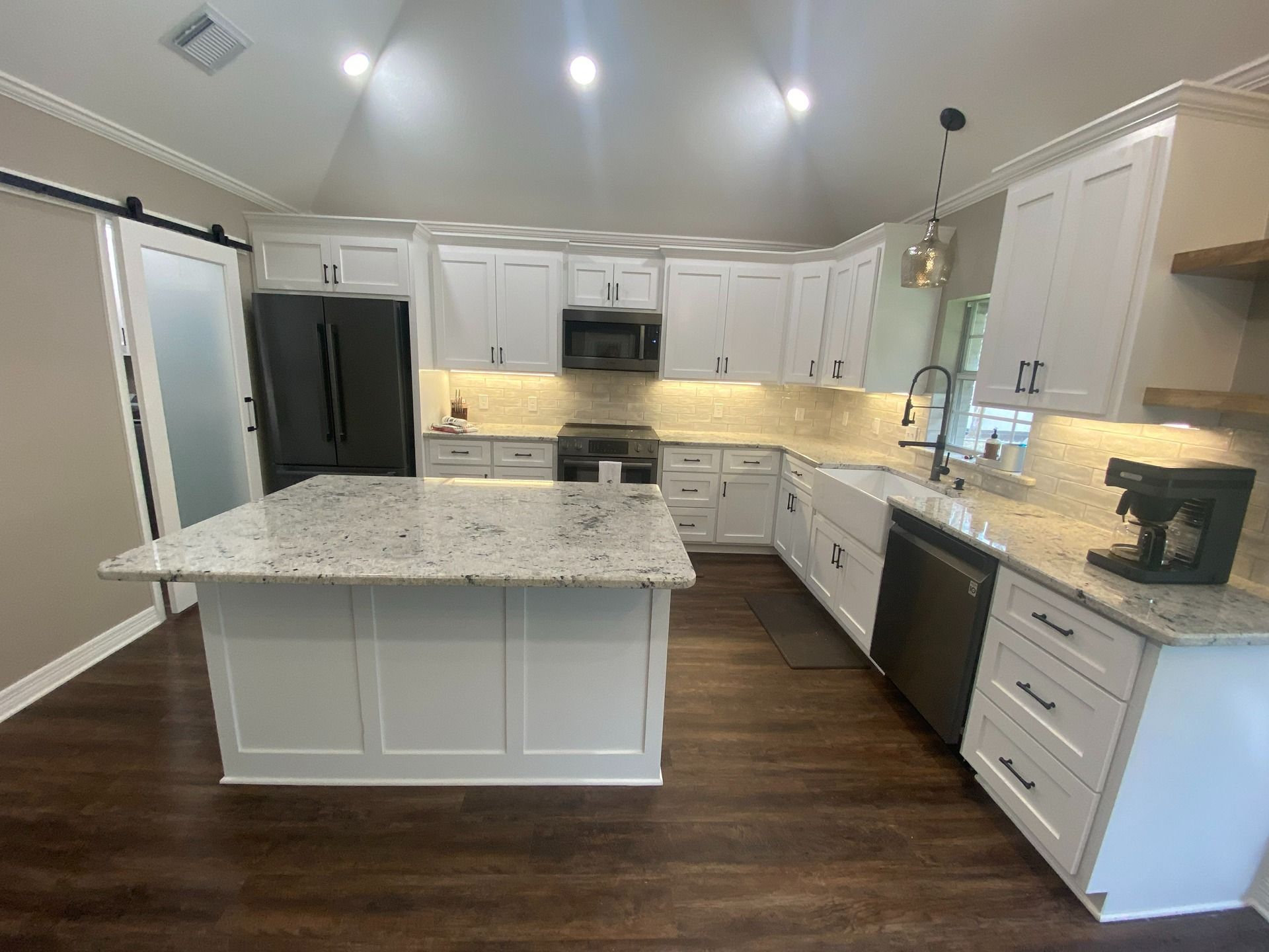 A kitchen with white cabinets, a large granite island, dark wood floors, and a dark refrigerator under vaulted ceilings.