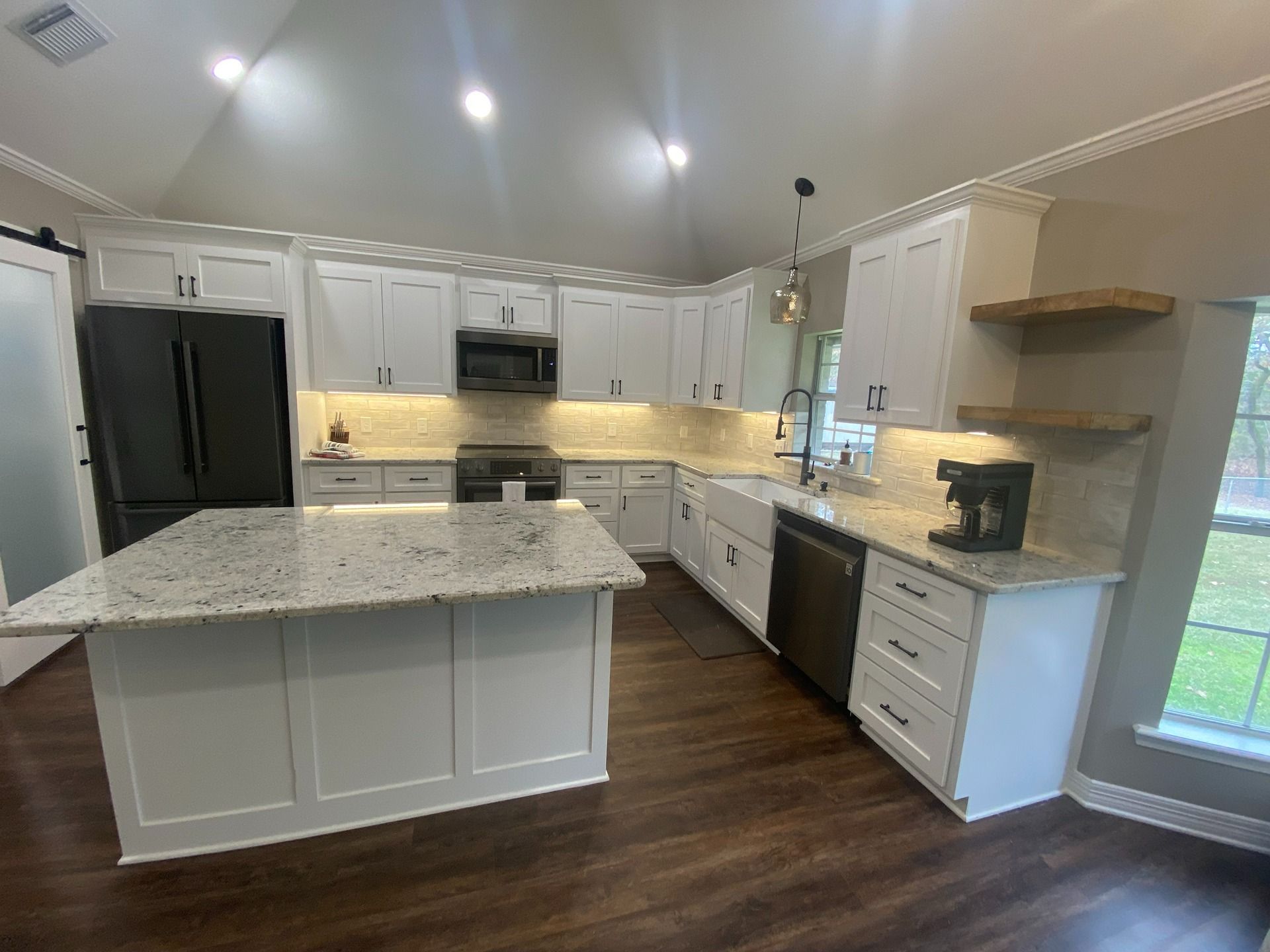 A modern kitchen with white cabinets, granite island, dark wood floors, black refrigerator, and open wooden wall shelves.