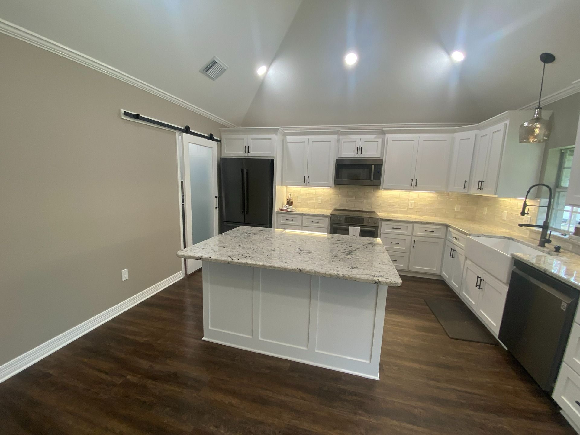 A modern kitchen with white cabinets, a granite-topped island, stainless appliances, and dark wood floors under a vaulted ceiling.