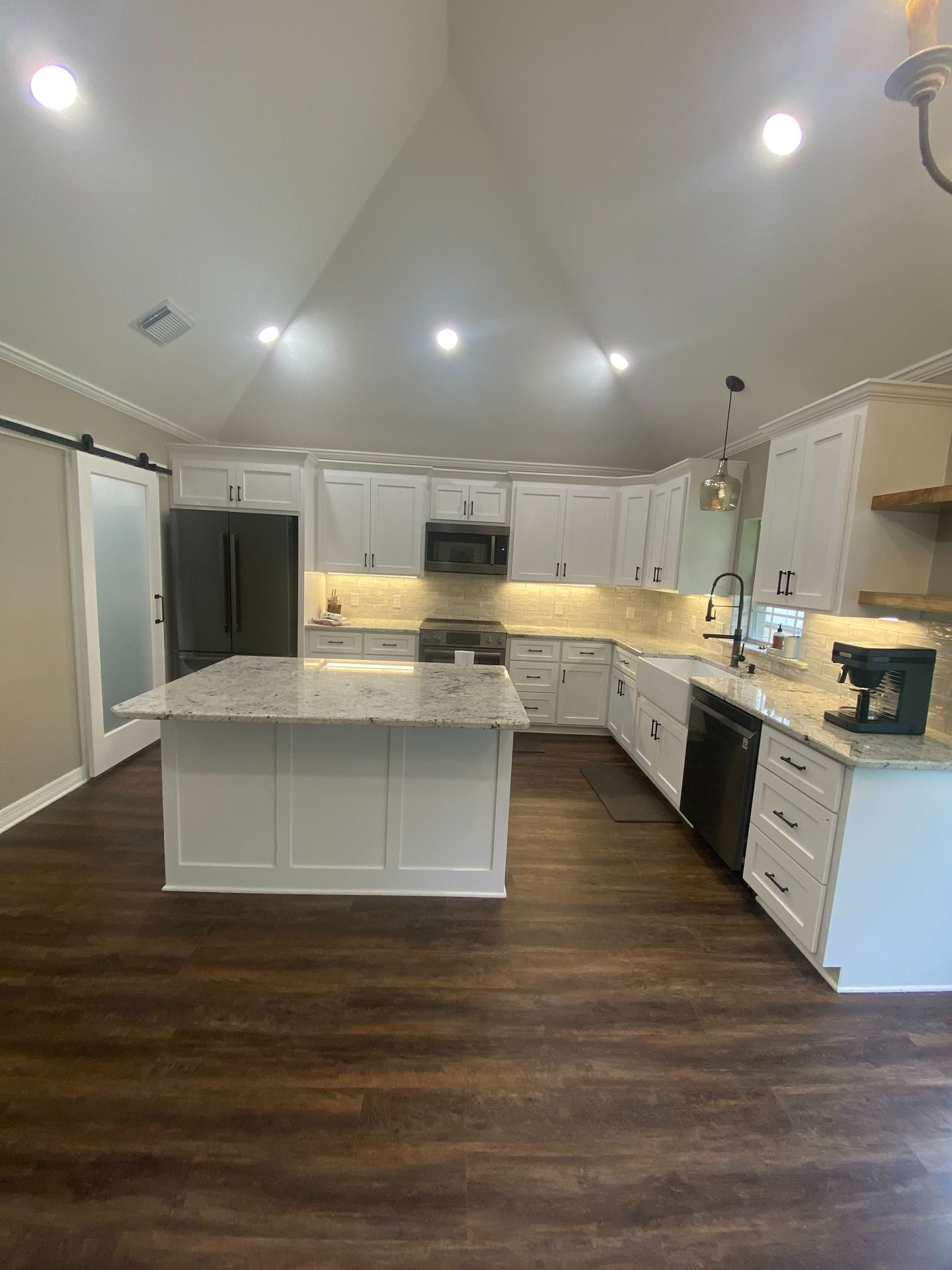 A modern kitchen with white cabinets, a granite island, dark flooring, a vaulted ceiling, and stainless steel appliances.