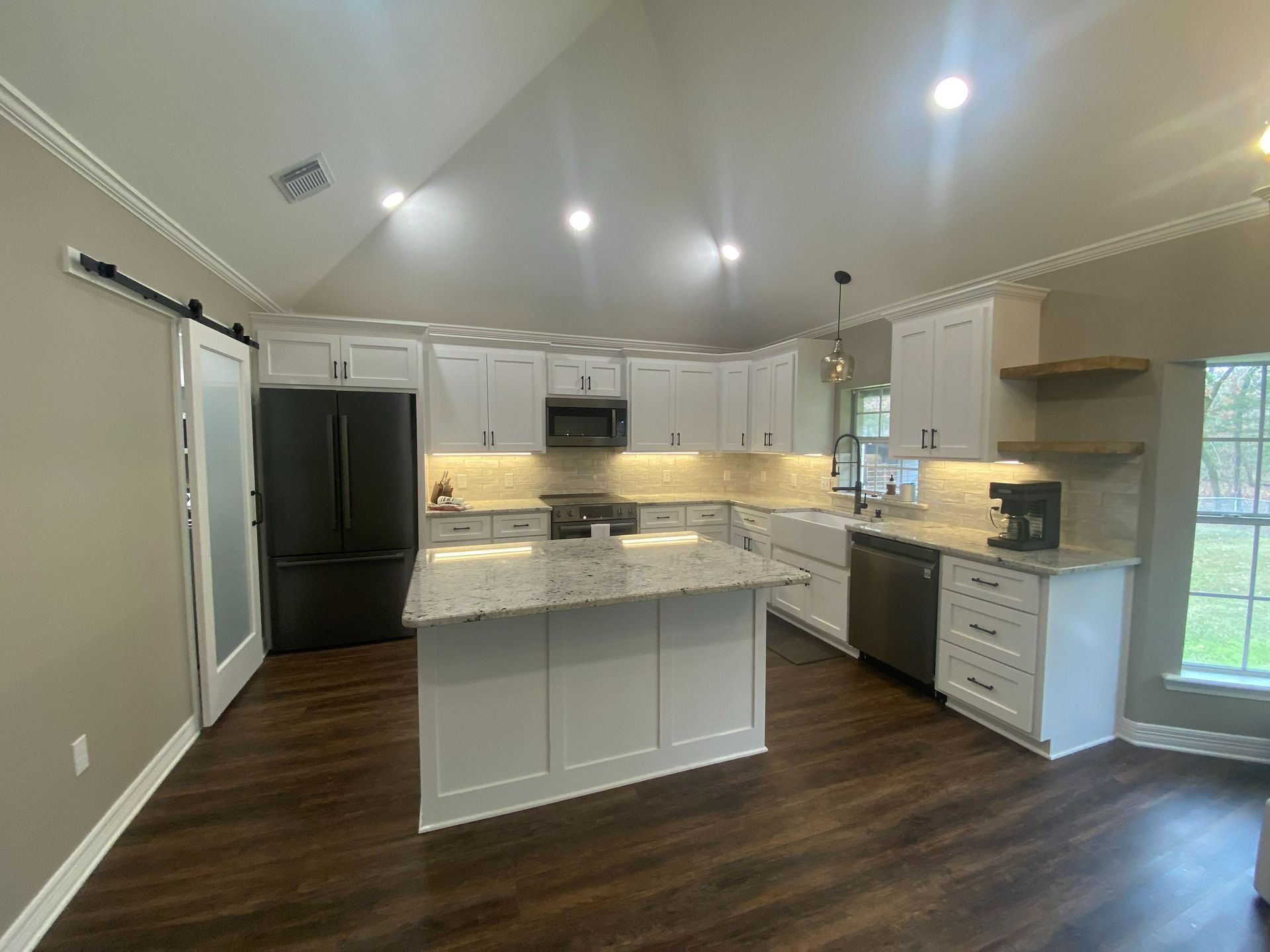 A modern kitchen with a white island, dark wood floors, white cabinets, black appliances, and floating wood shelves.