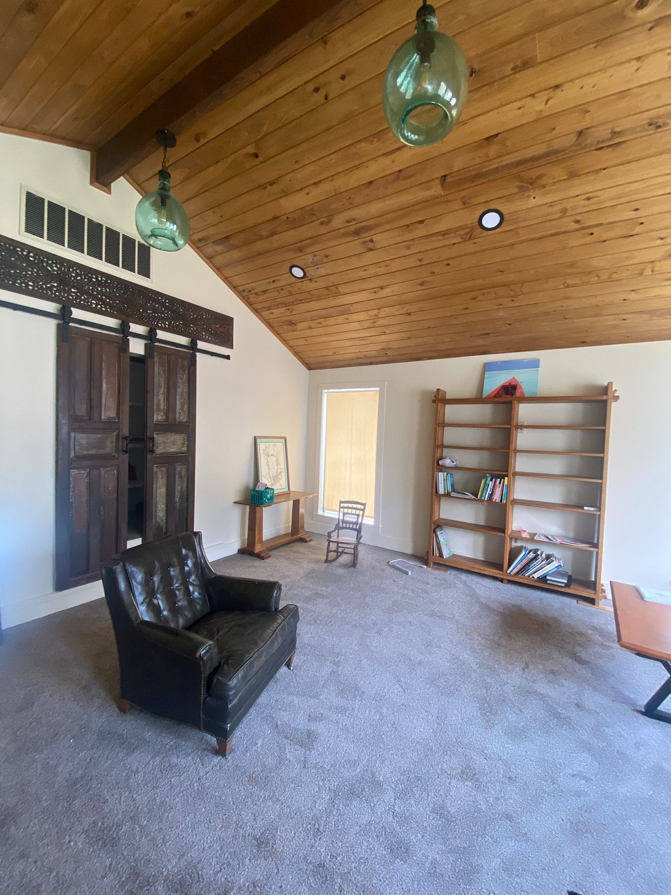 A living room featuring a wood-vaulted ceiling, glass pendant lights, a dark leather armchair, and wooden bookshelves.