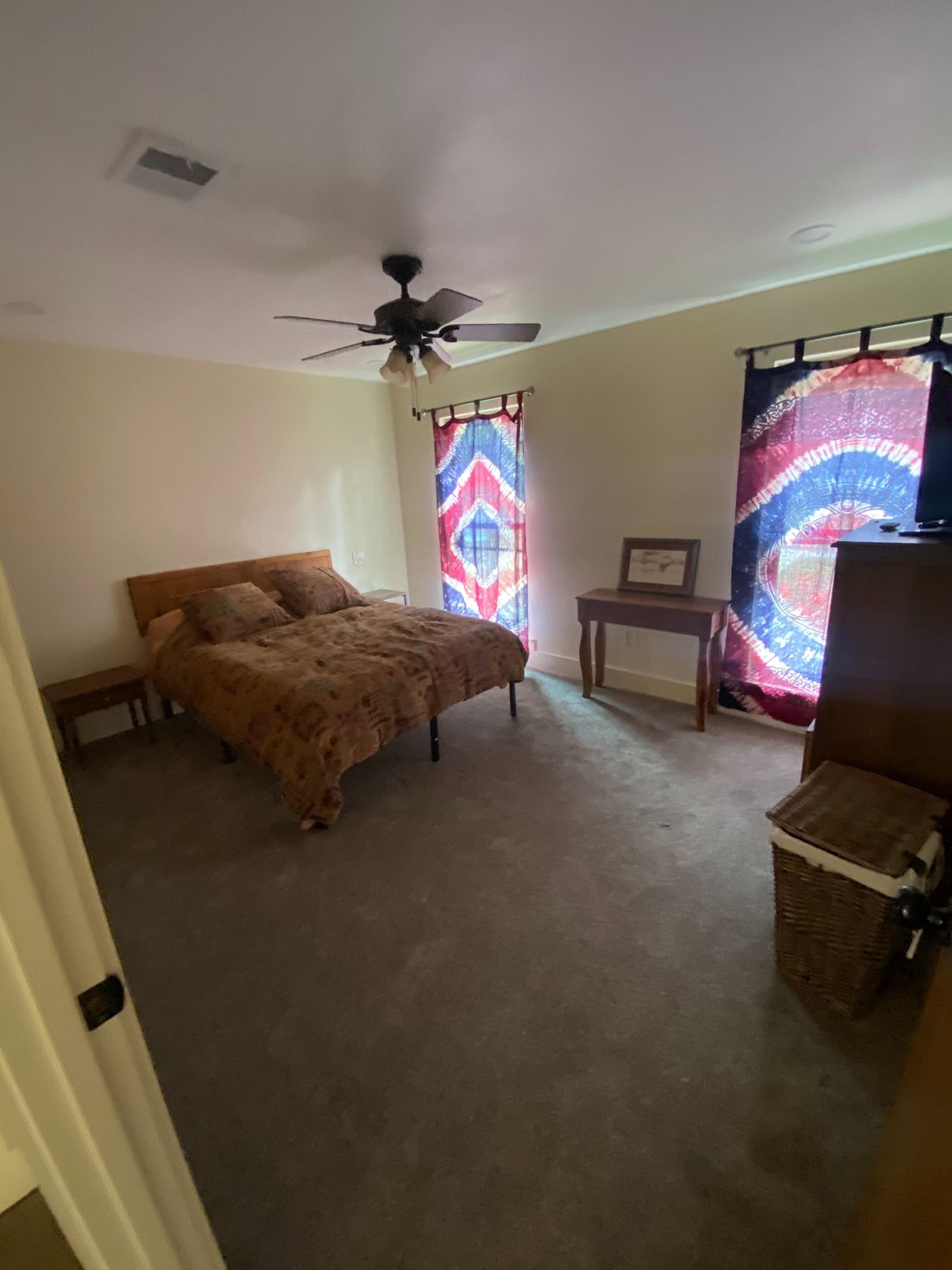 A bedroom with a brown bed, a ceiling fan, a small wooden desk, and large windows with red, white, and blue patterned drapes.