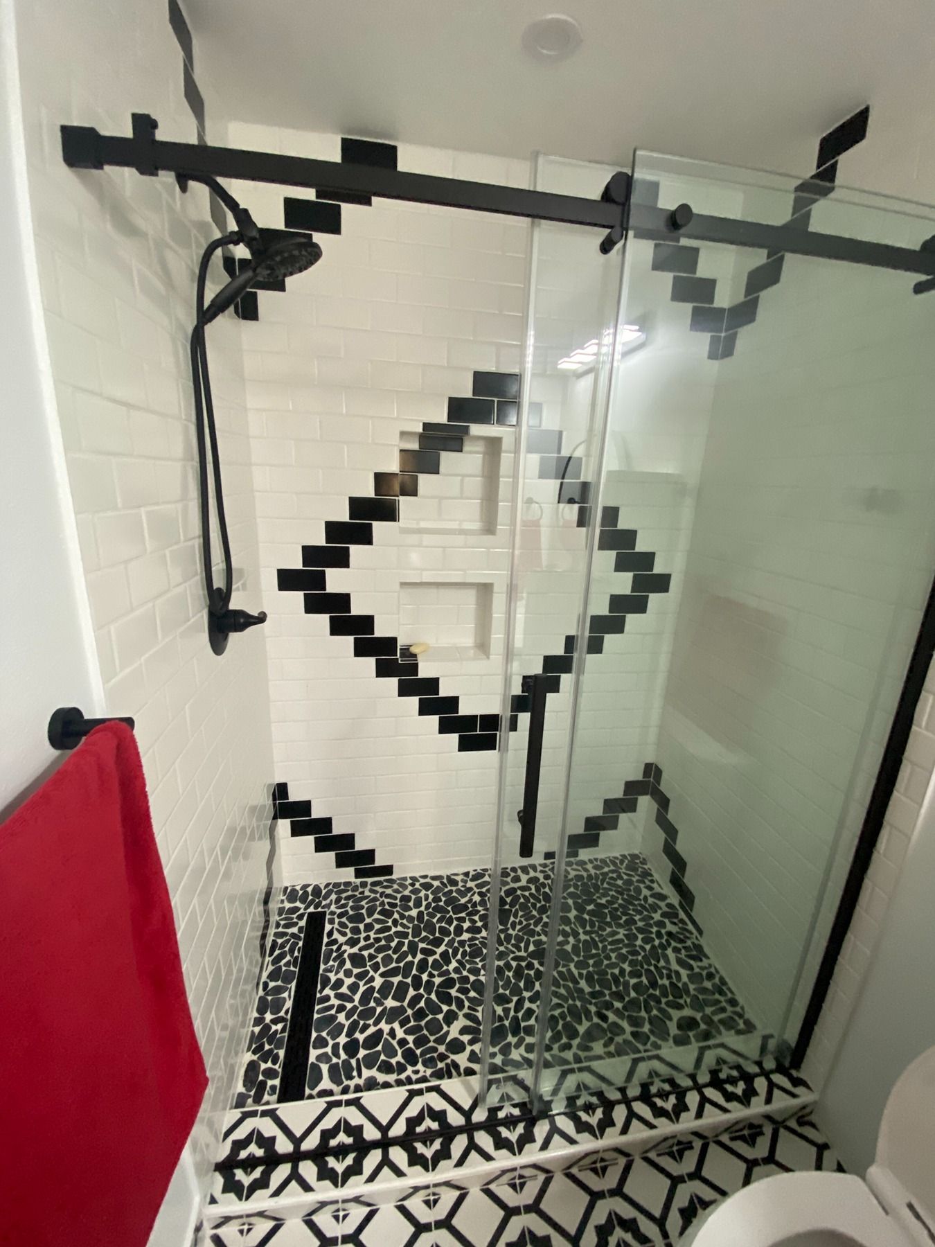 Modern shower with white subway tiles, a black diamond geometric pattern, pebble floor, and glass sliding door.