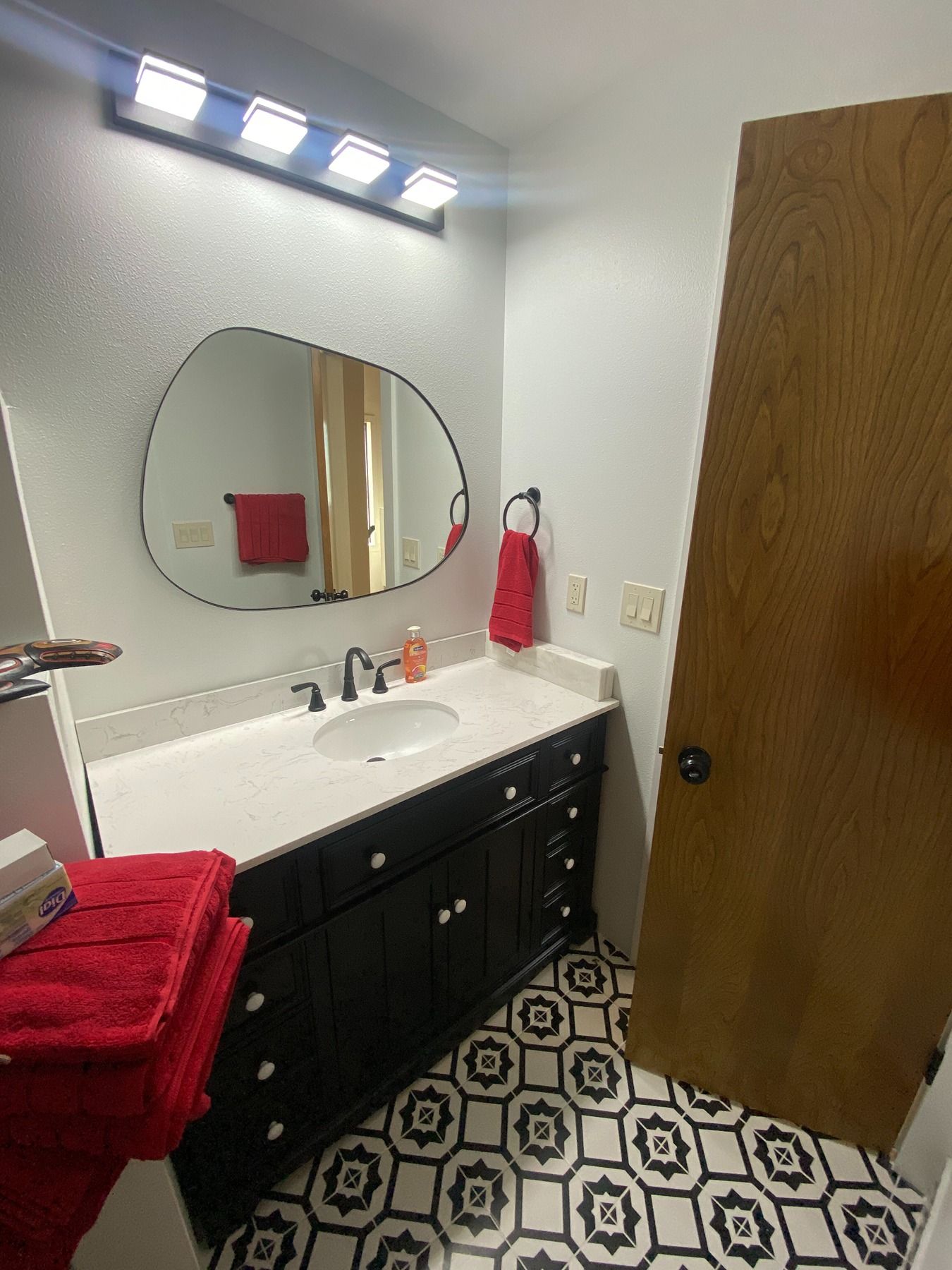 A bathroom vanity with a dark cabinet, white countertop, irregular-shaped mirror, and patterned floor tile.
