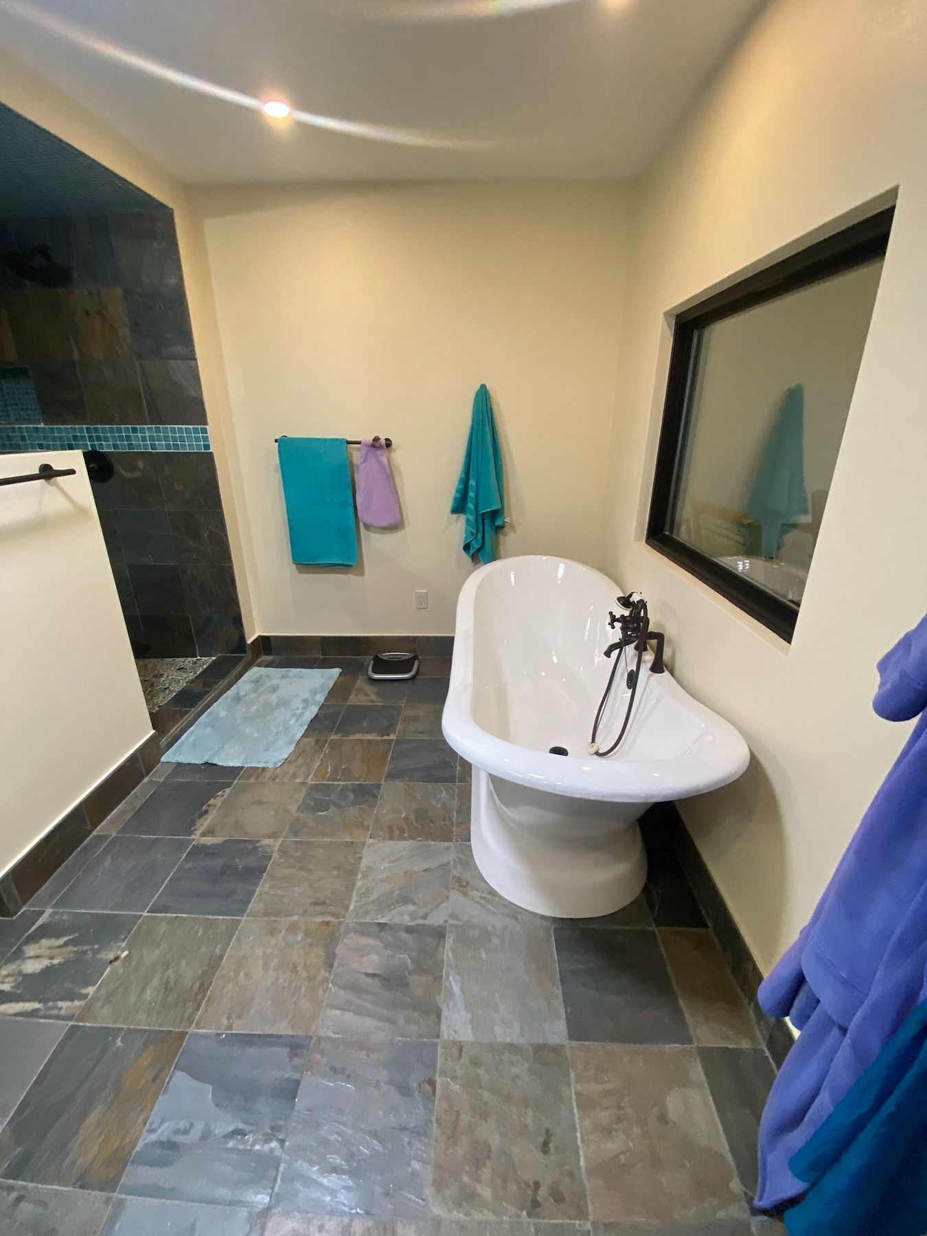 A modern bathroom featuring a white freestanding bathtub on slate-tiled floors, with towels hanging on the light-toned walls.