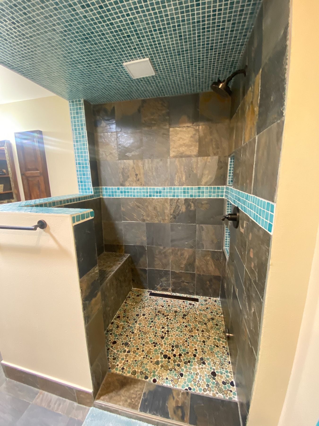A walk-in shower with dark slate wall tiles, a built-in bench, turquoise mosaic accents, and a pebble-stone floor.