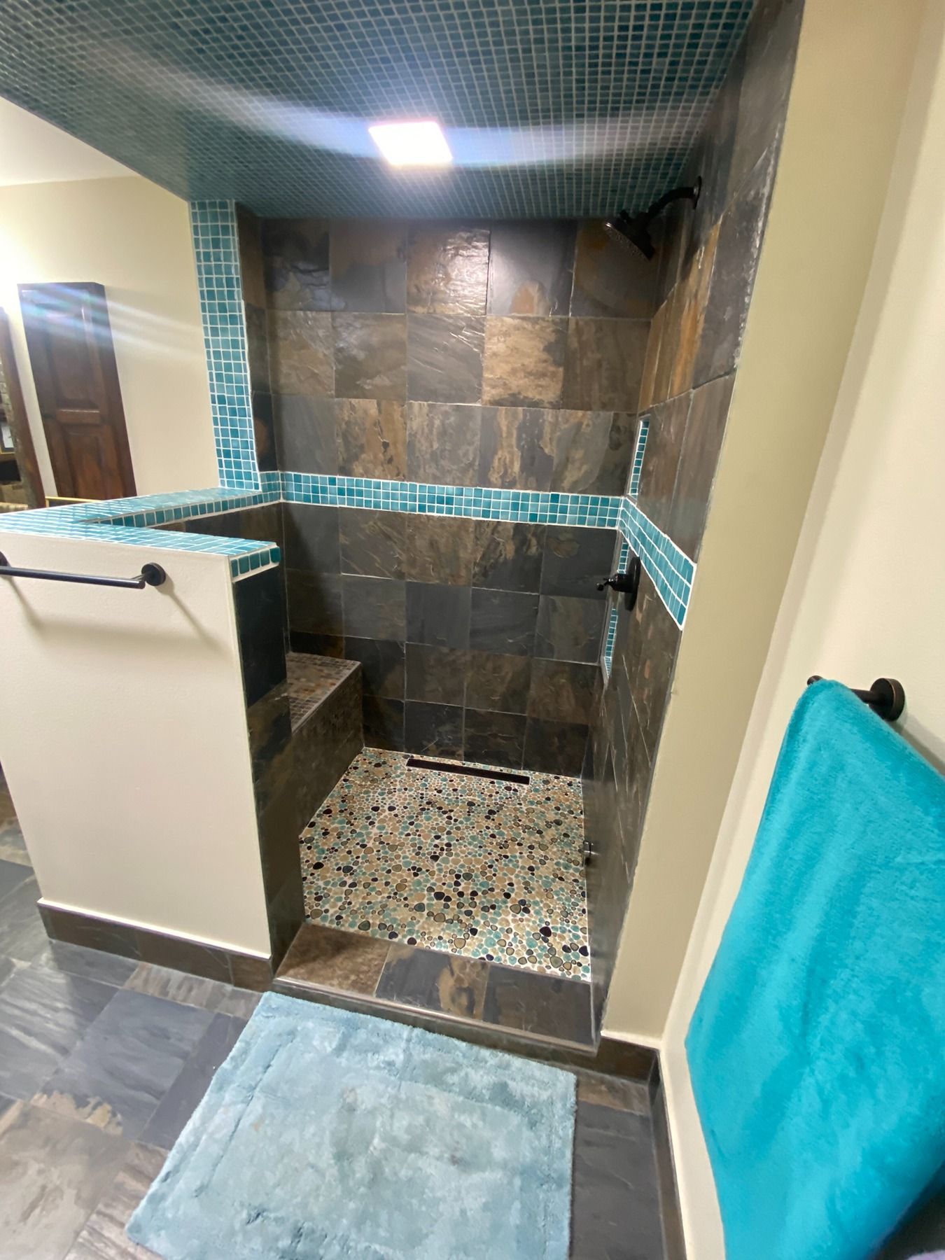 A tiled walk-in shower with dark brown stone walls, a pebble floor, and teal accent tiles, featuring a built-in bench.