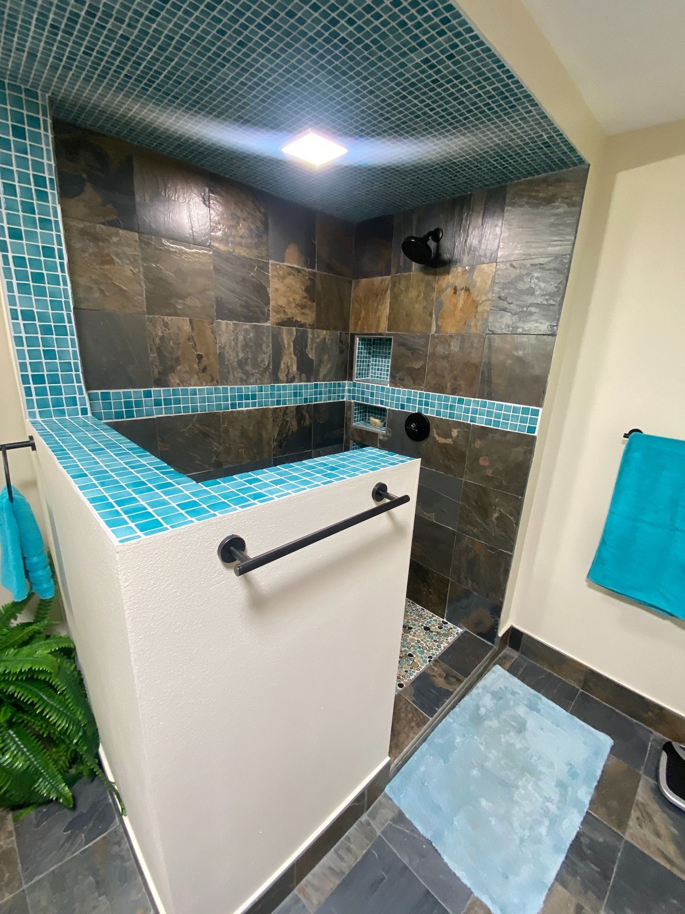 A walk-in shower with dark slate wall tiles and vibrant turquoise mosaic accents on the ceiling and half-wall.