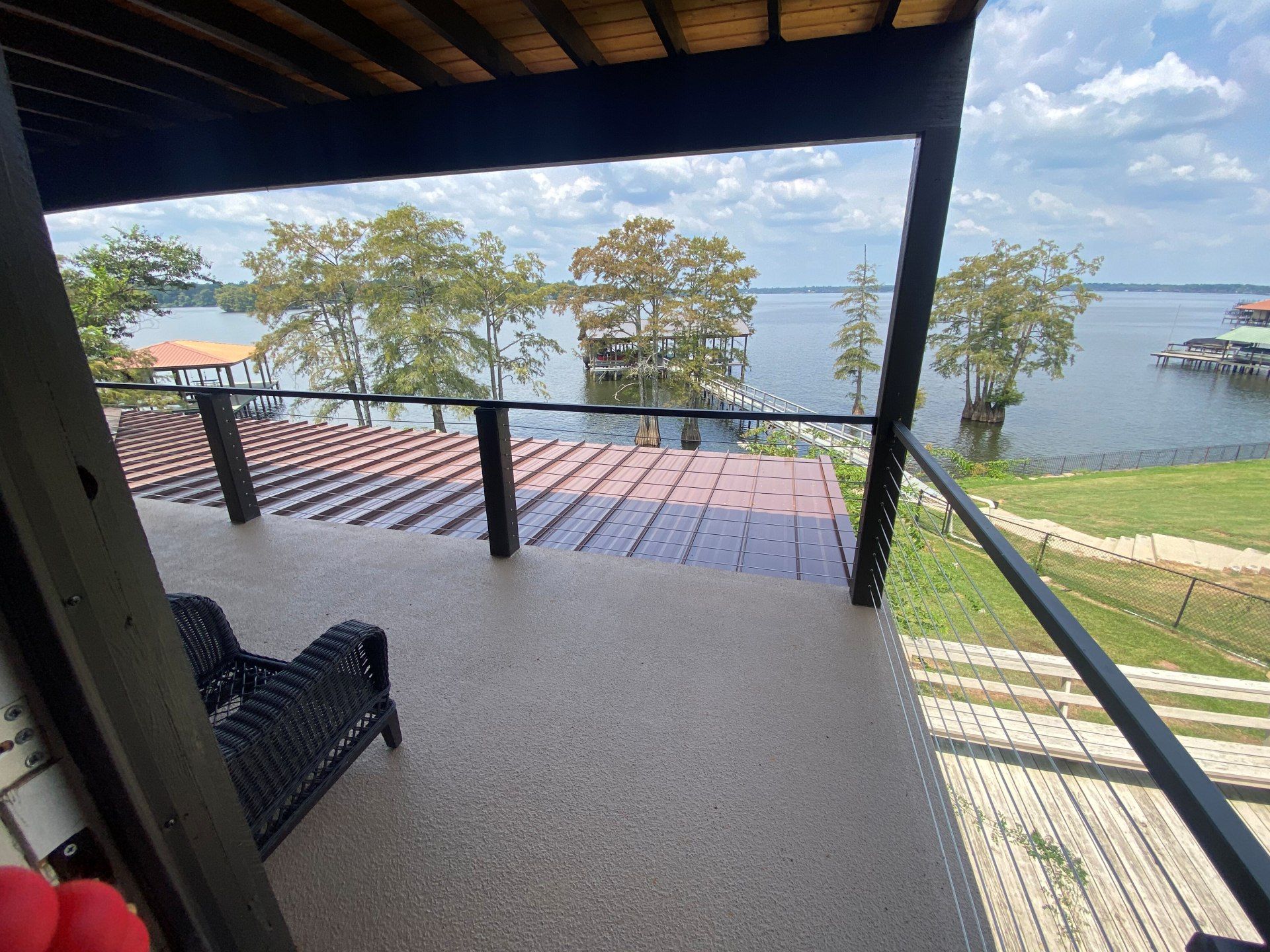 A balcony view overlooking a calm lake, with trees in the water, a dark roof below, and a black metal railing.