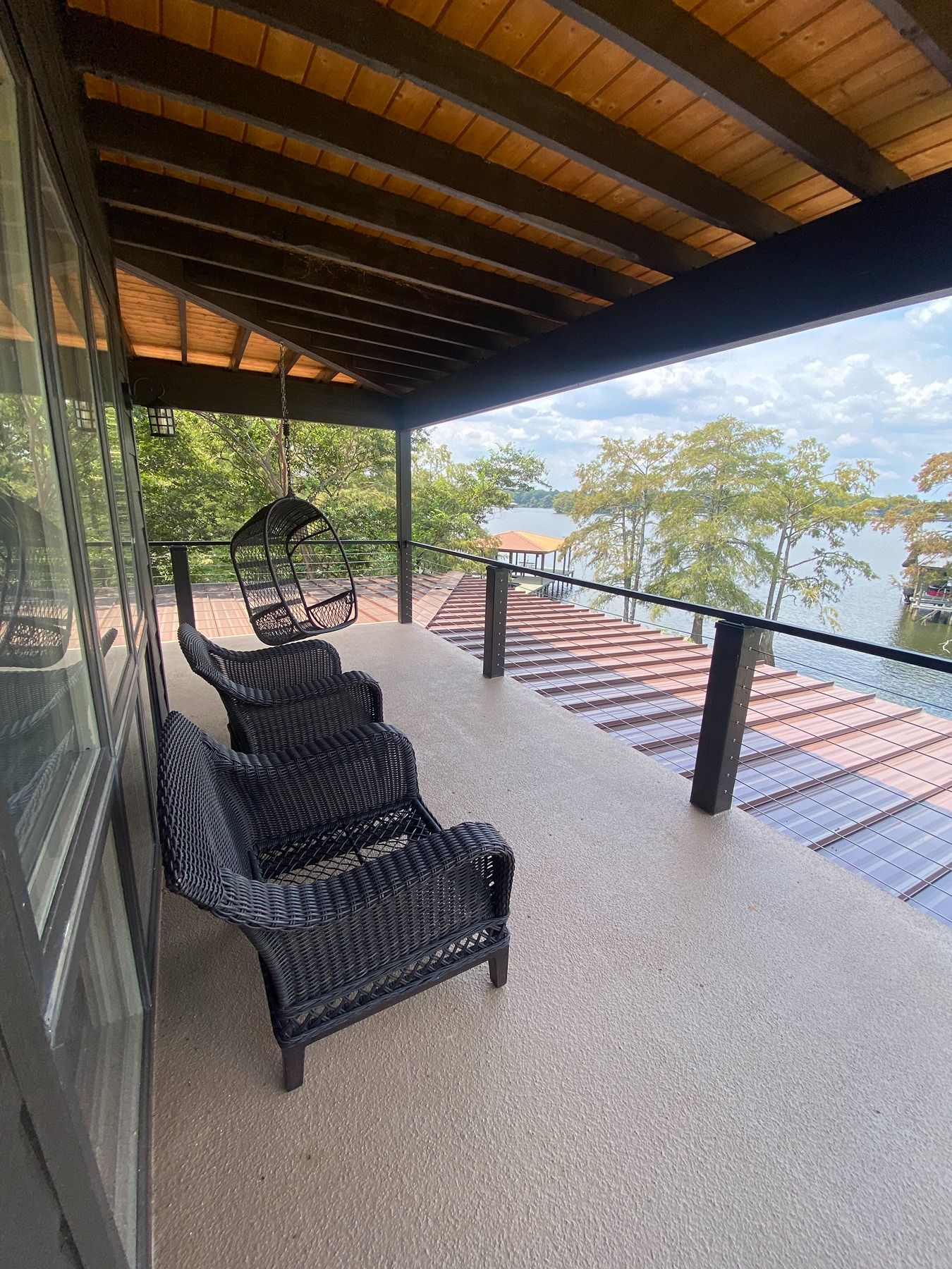 A balcony with dark wicker seating overlooking a lake and trees under a cloudy sky.