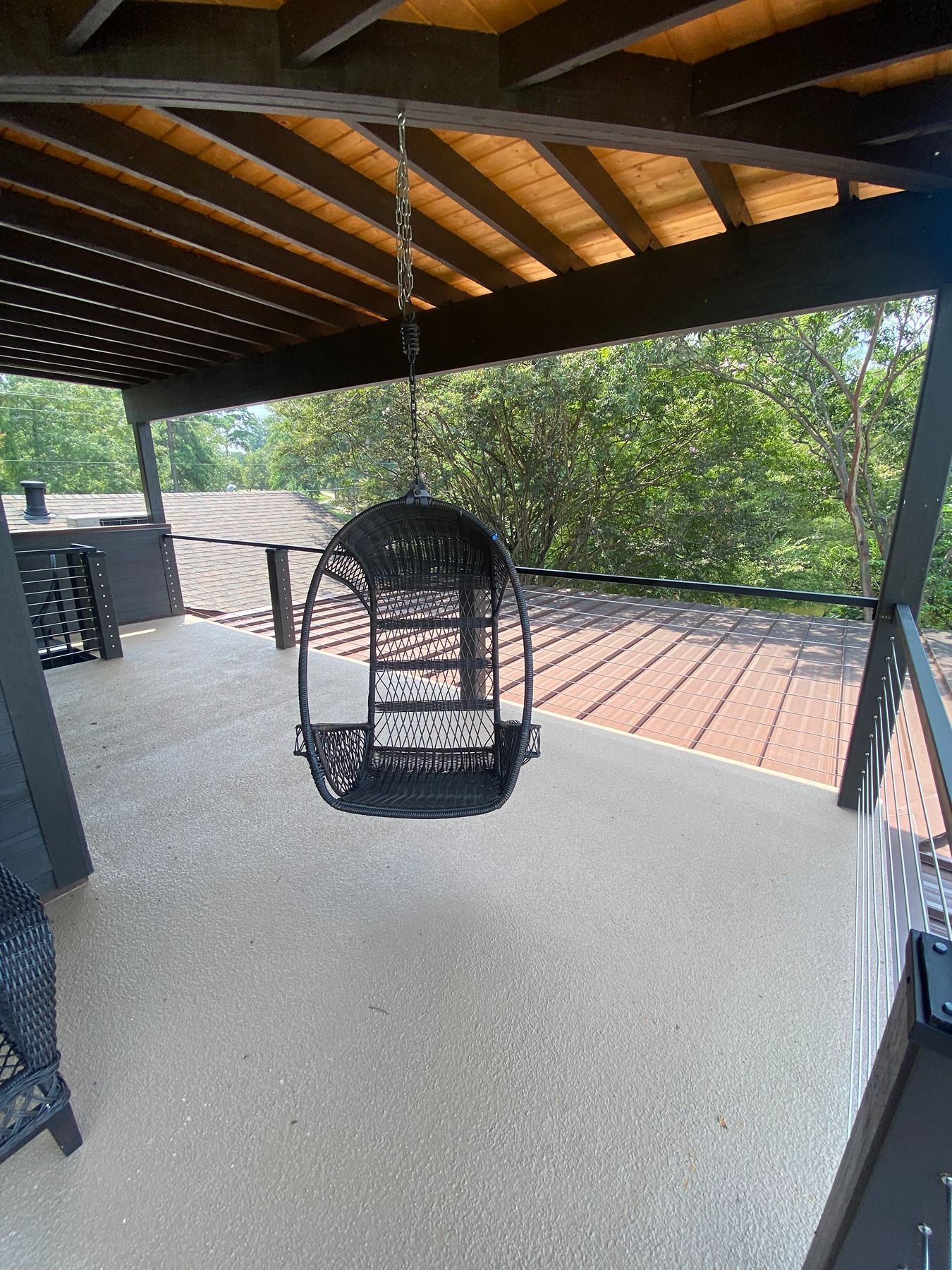 A black, egg-shaped hanging chair suspended from a covered patio ceiling overlooking a treed landscape.