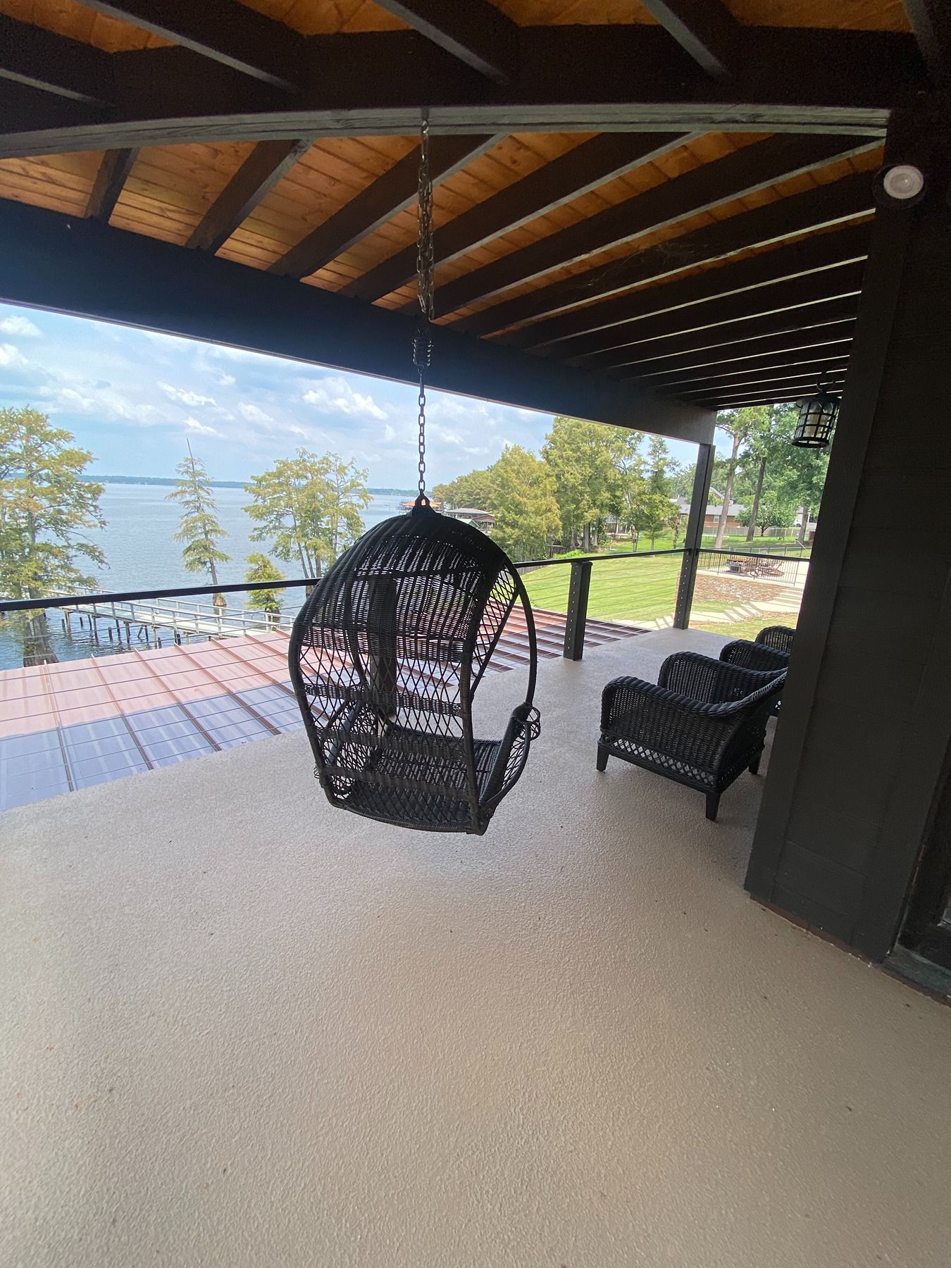 A black hanging basket chair on a covered patio overlooking a lake, with additional outdoor seating nearby.