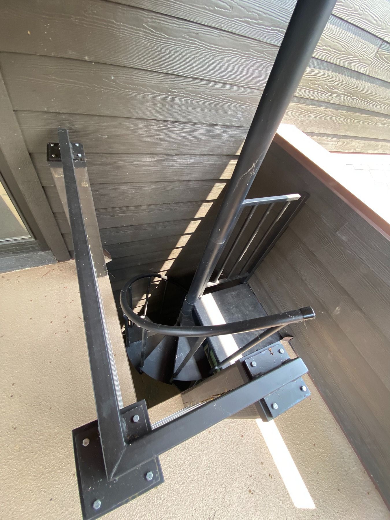 A black metal spiral staircase viewed from above, installed on a deck with dark wood siding.