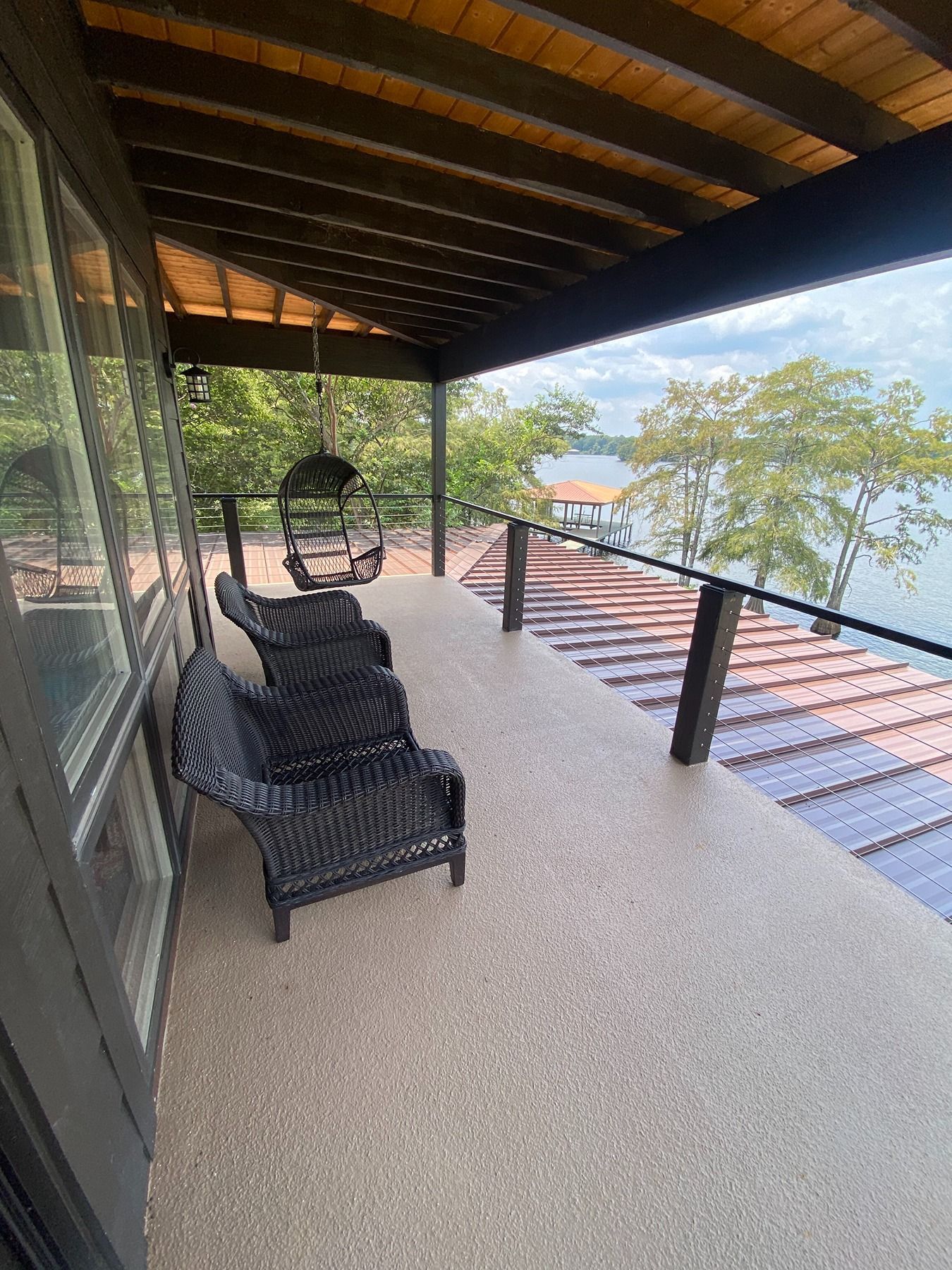 Covered balcony with wicker chairs and a hanging egg chair overlooking a lake on a sunny day.
