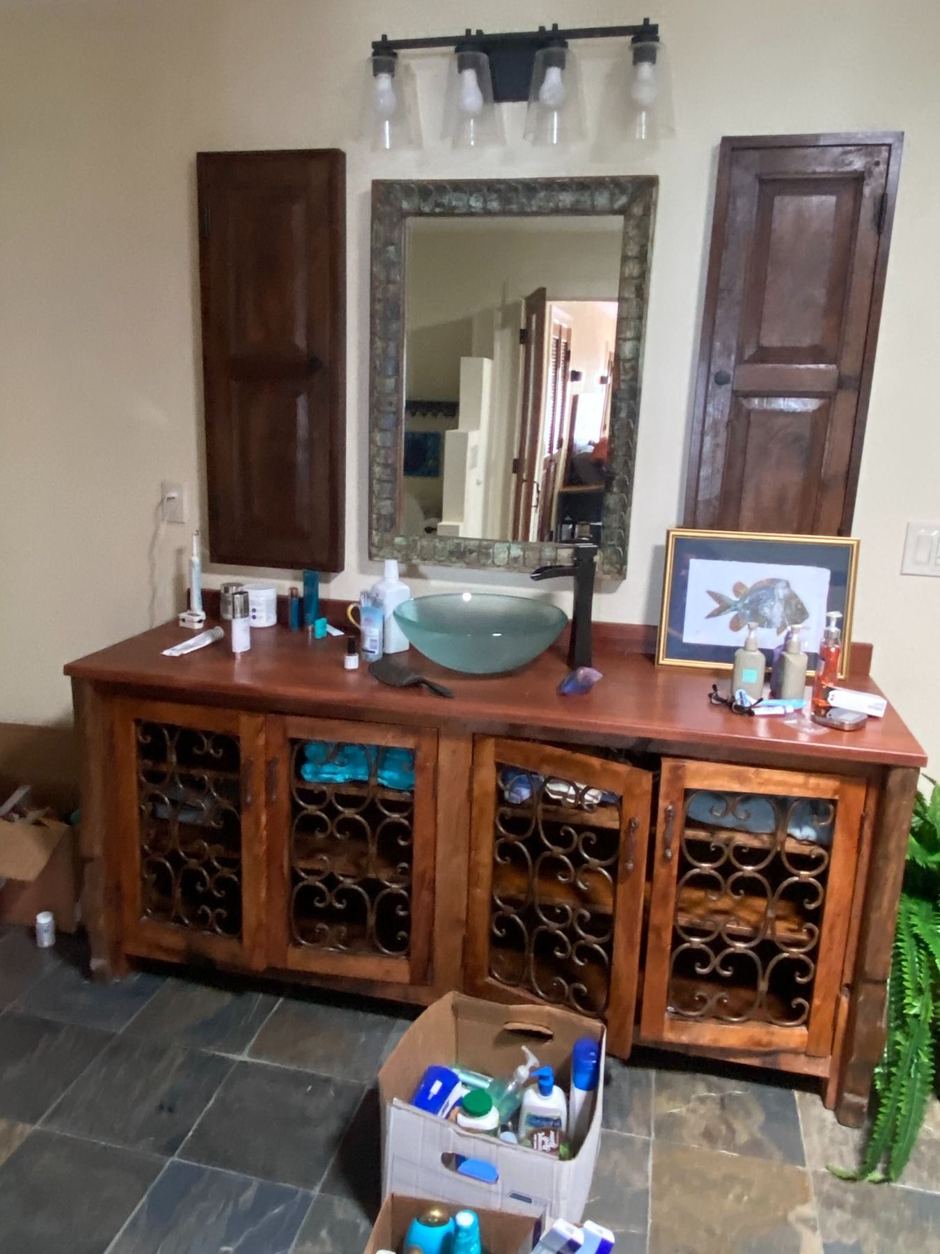 A bathroom vanity with a wood frame, glass vessel sink, ornate metal cabinet doors, and wall-mounted decor in a tiled room.