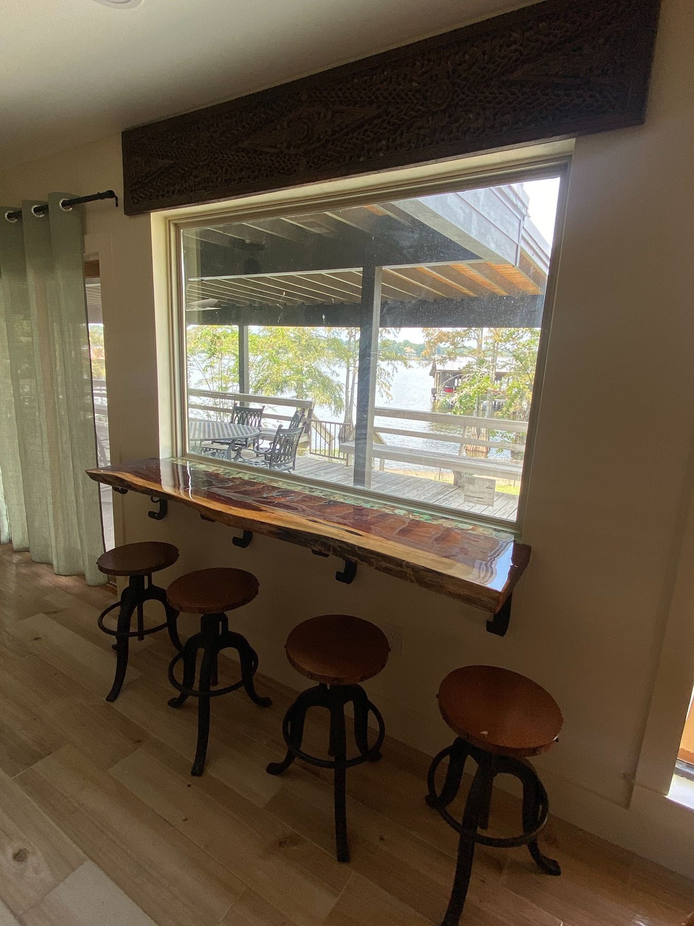A live-edge wood bar top with four metal swivel stools sits beneath a large window overlooking a lake and deck.