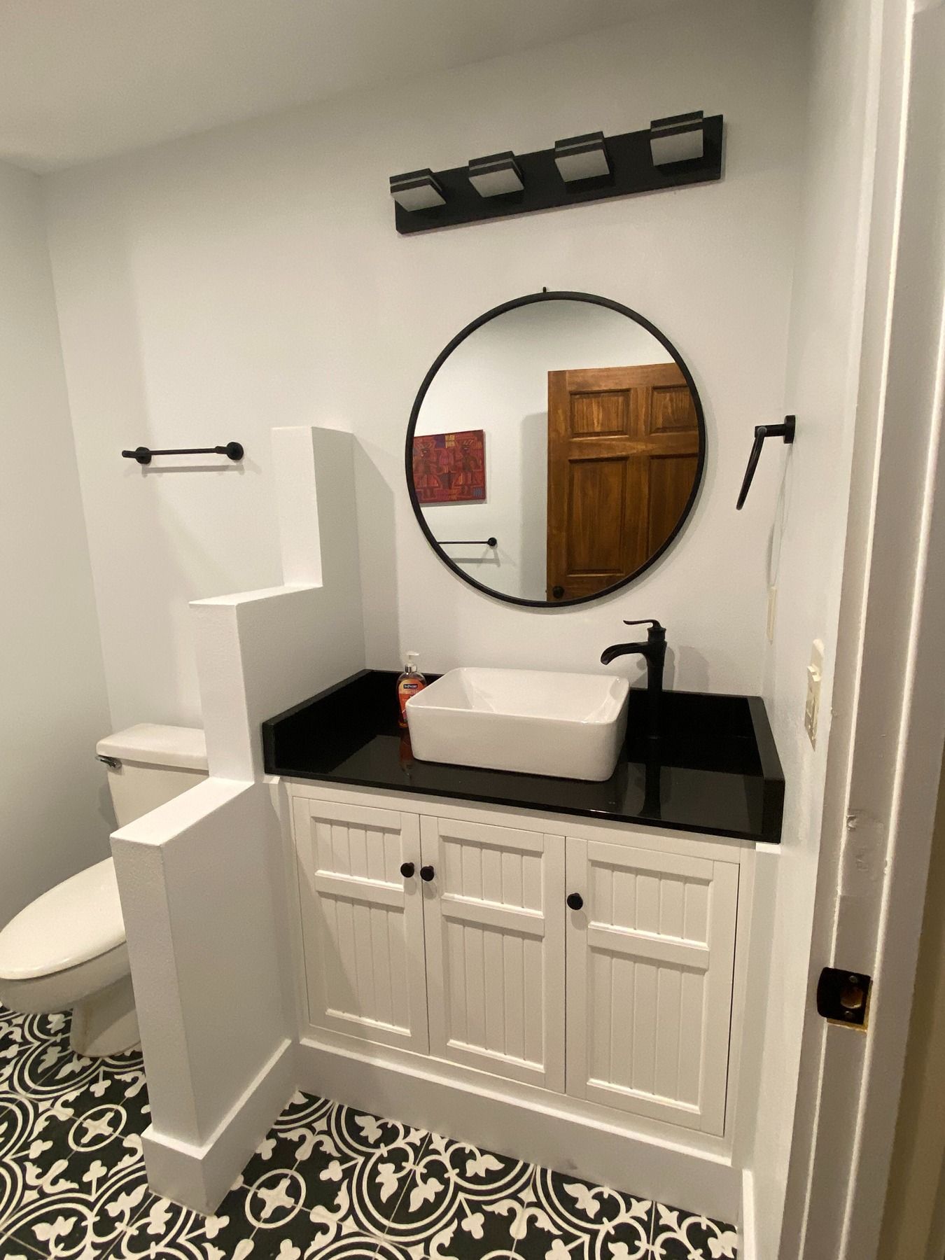 A bathroom with a white vanity, black countertop, square vessel sink, round mirror, and patterned black-and-white floor.
